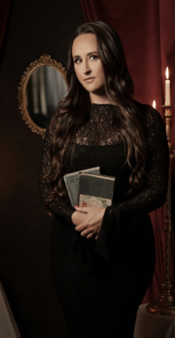 Nikki Bodie in black lace, holding old books beside a candle, a gilded mirror behind her