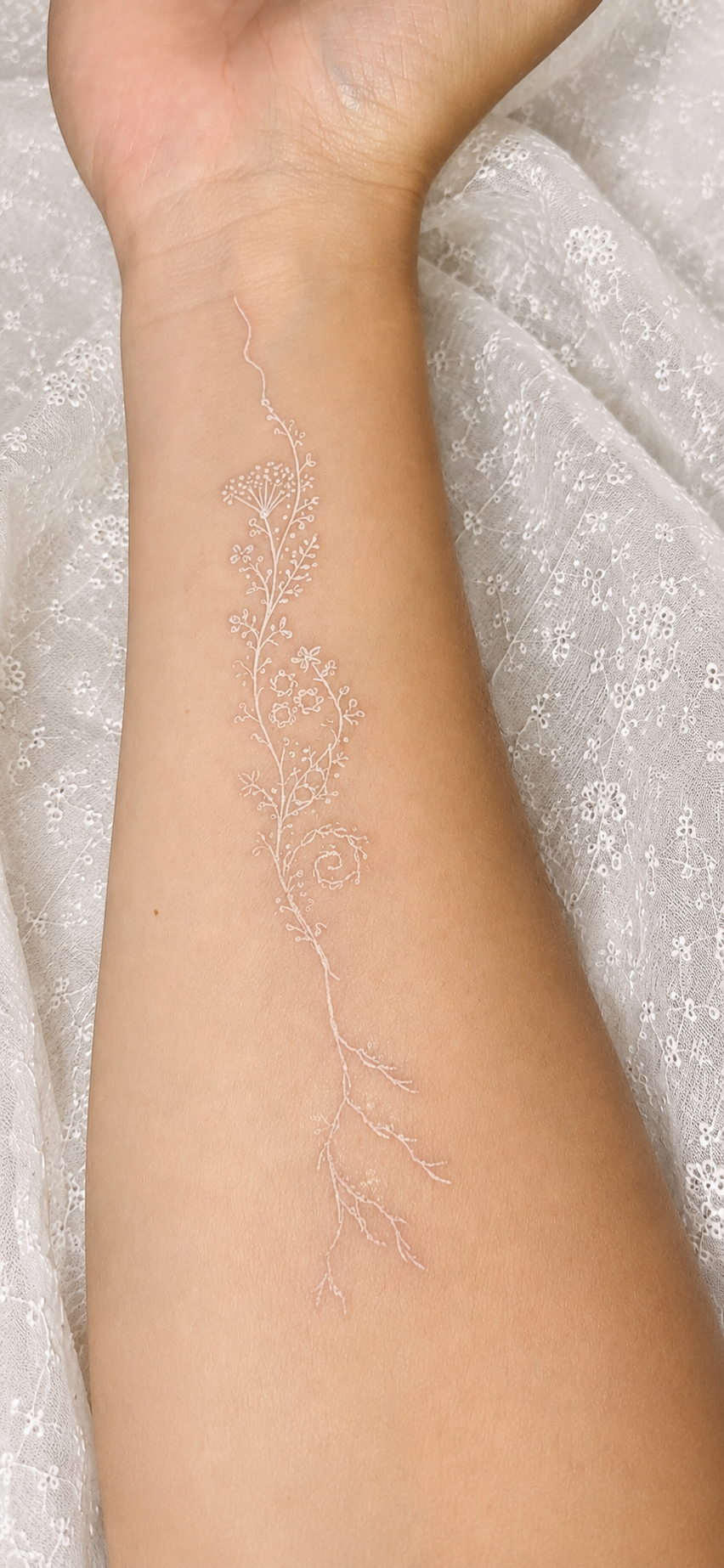 A delicate white-ink tattoo of a flowering vine along an inner forearm, laid against white lace.