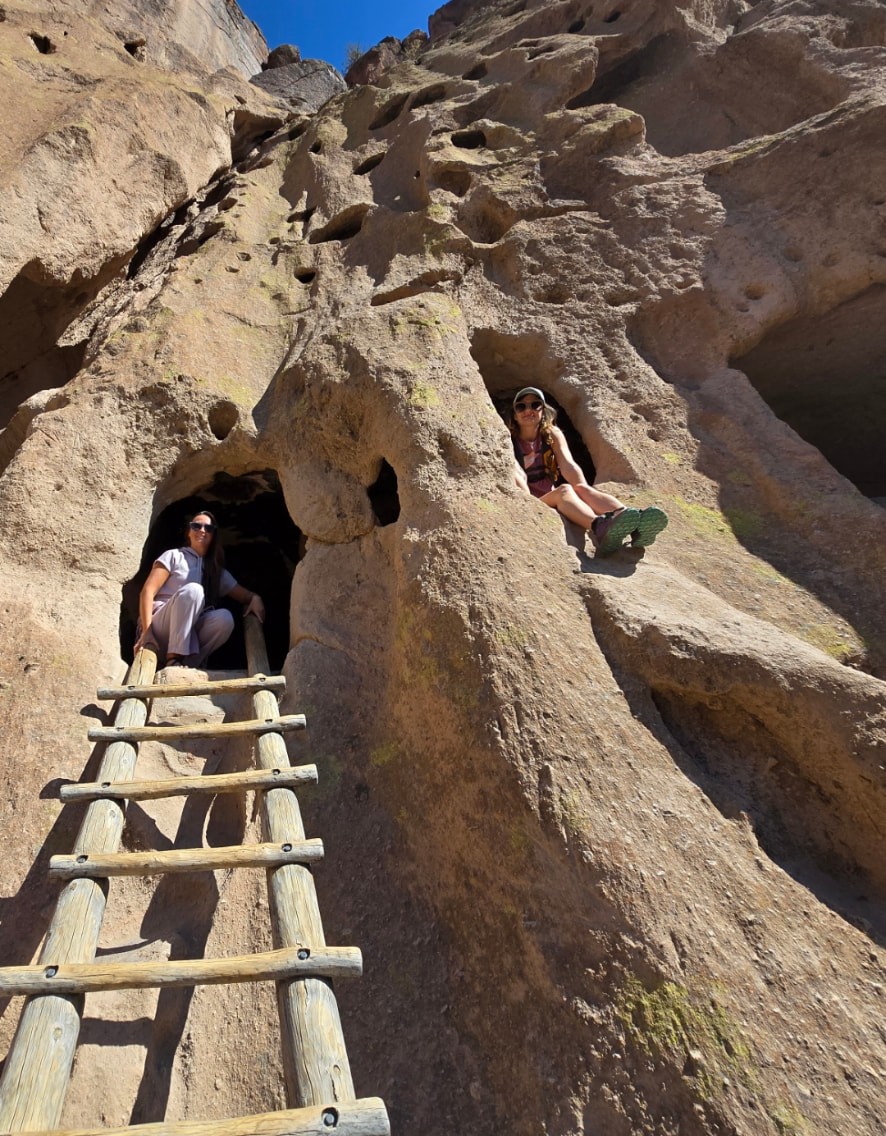 Nikki and Ali sitting in separate cliff dwelling openings above a wooden ladder.