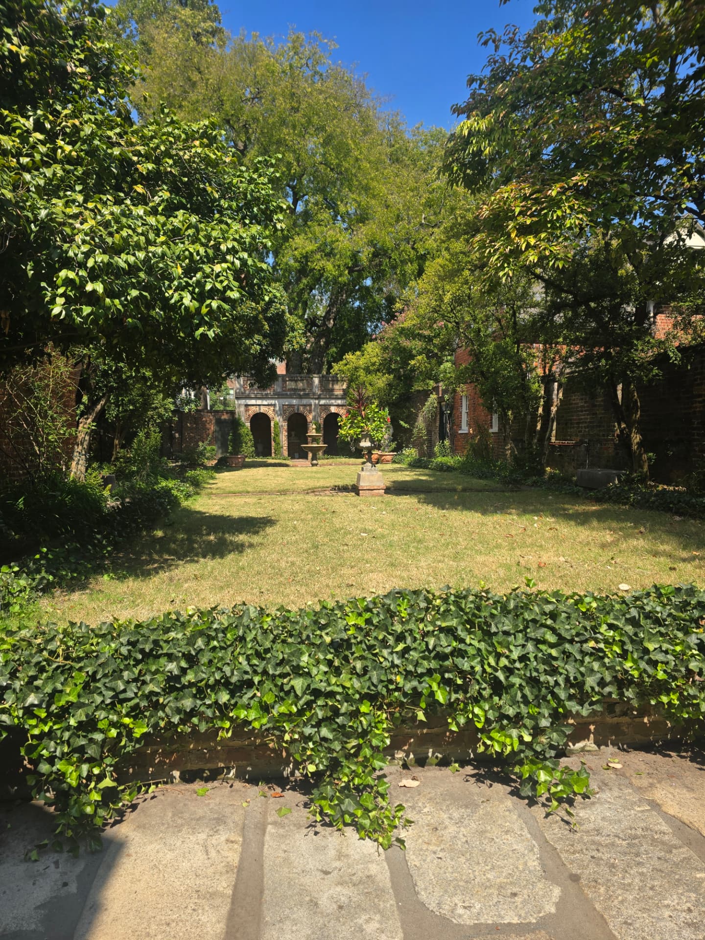 The Poe Museum's Enchanted Garden: a sunlit lawn framed by trees and brick walls, with a small fountain and the arched brick shrine at the far end.