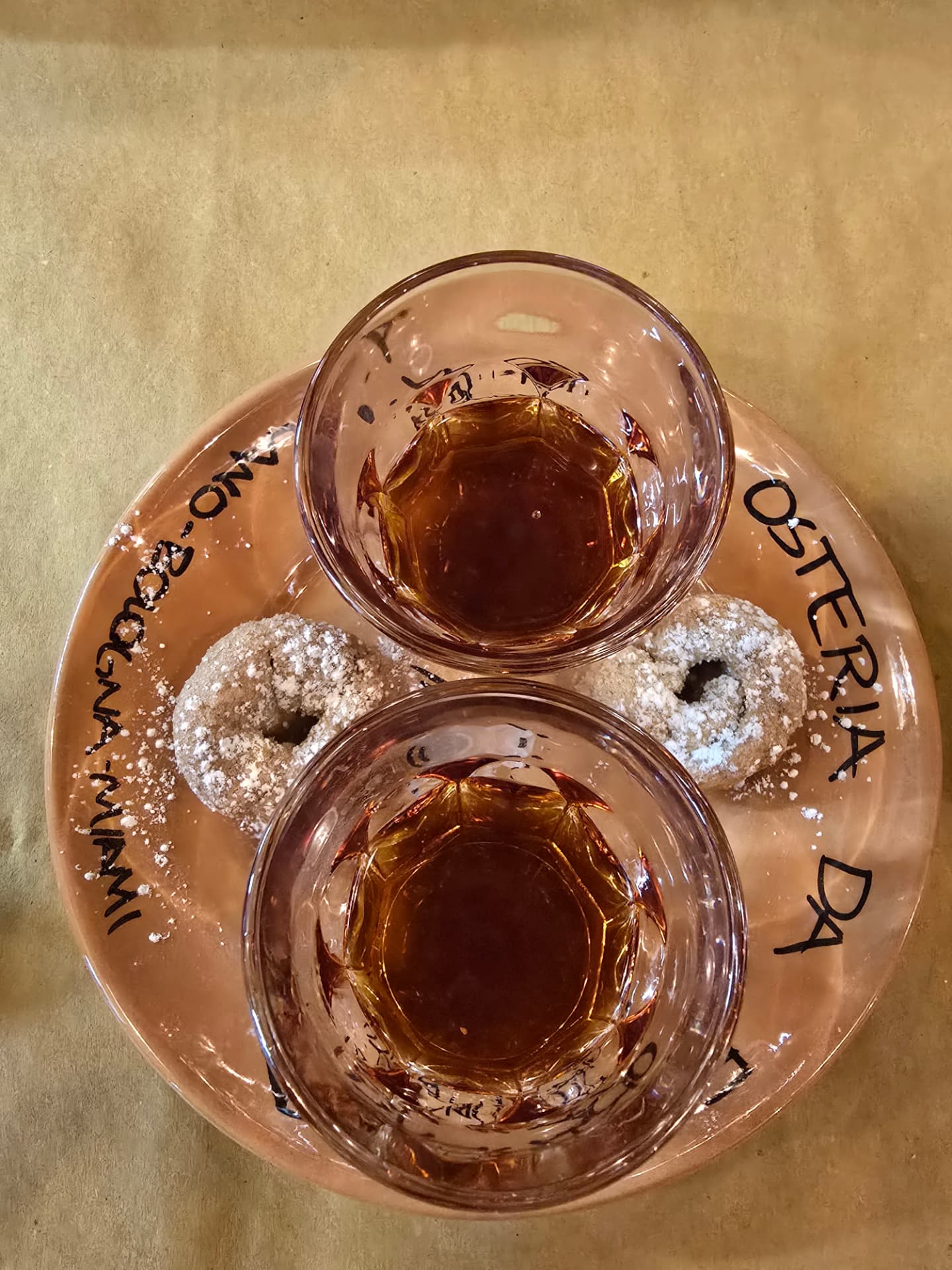 Two glasses of amber liqueur with sugared biscuits on a plate reading Osteria da, seen from above.