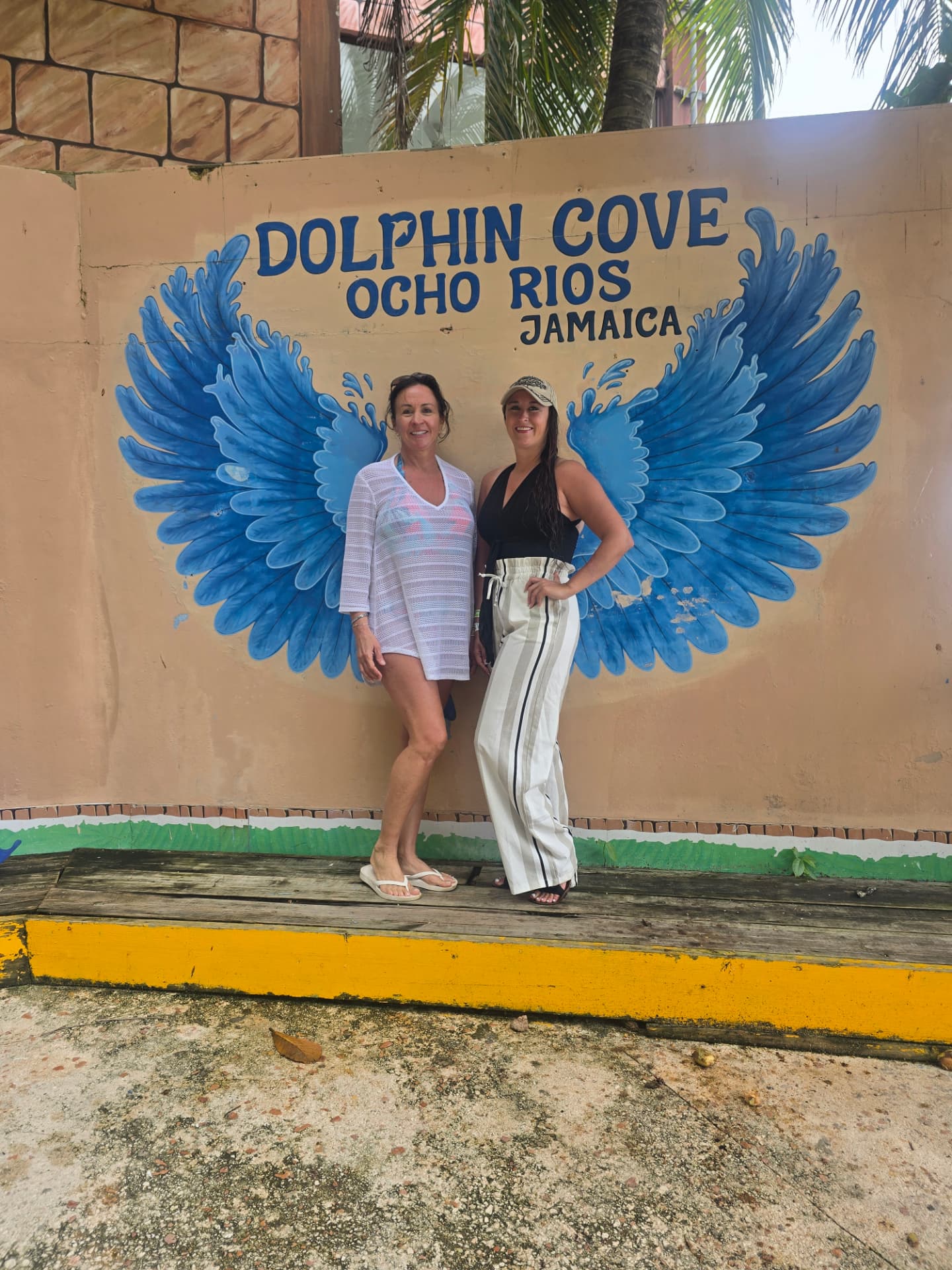 Nikki and her mom Patti standing in front of the blue painted wings of the Dolphin Cove Ocho Rios, Jamaica sign.