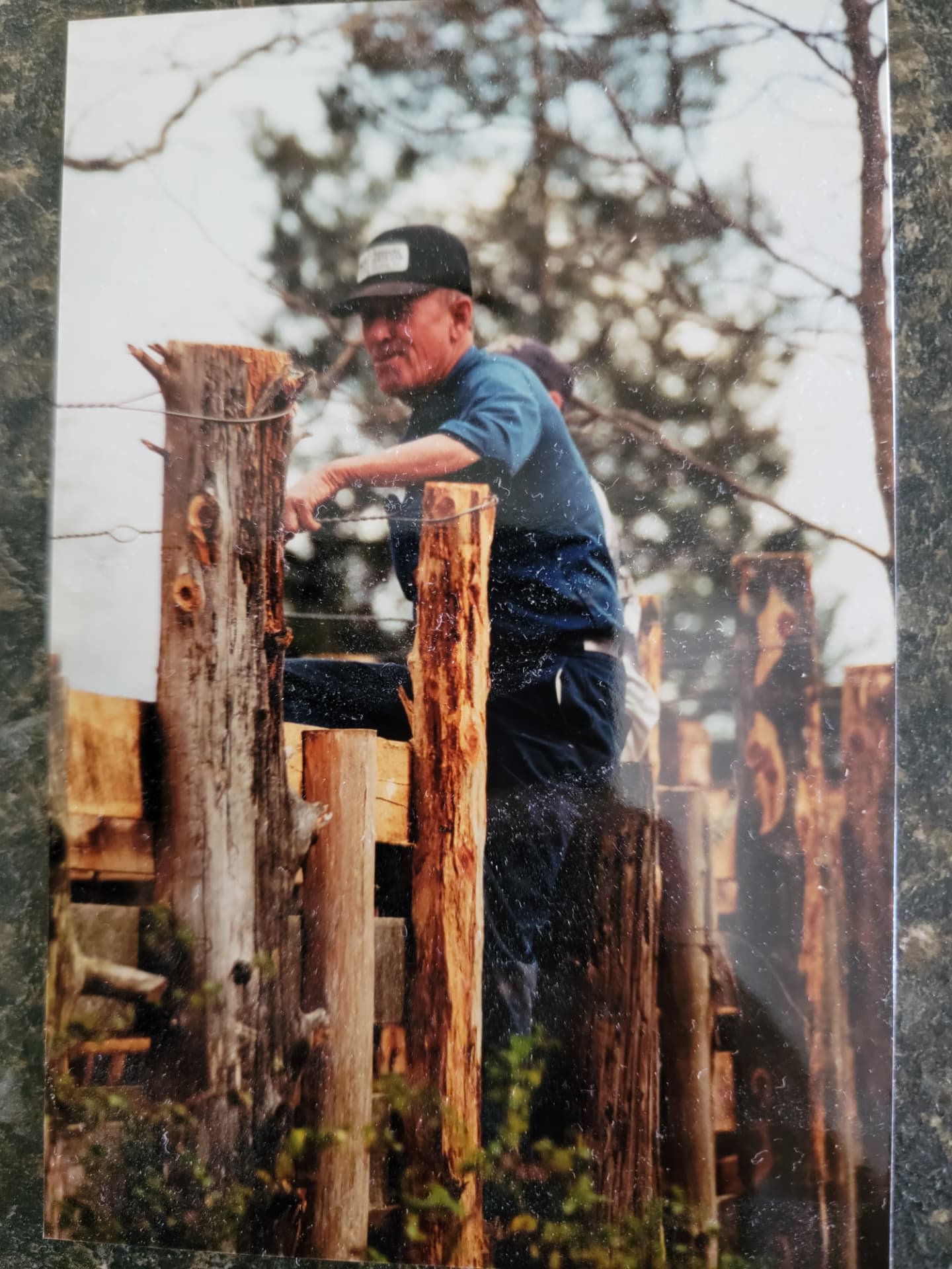 A faded color photograph of an older man in a black cap climbing over a weathered wooden post fence.