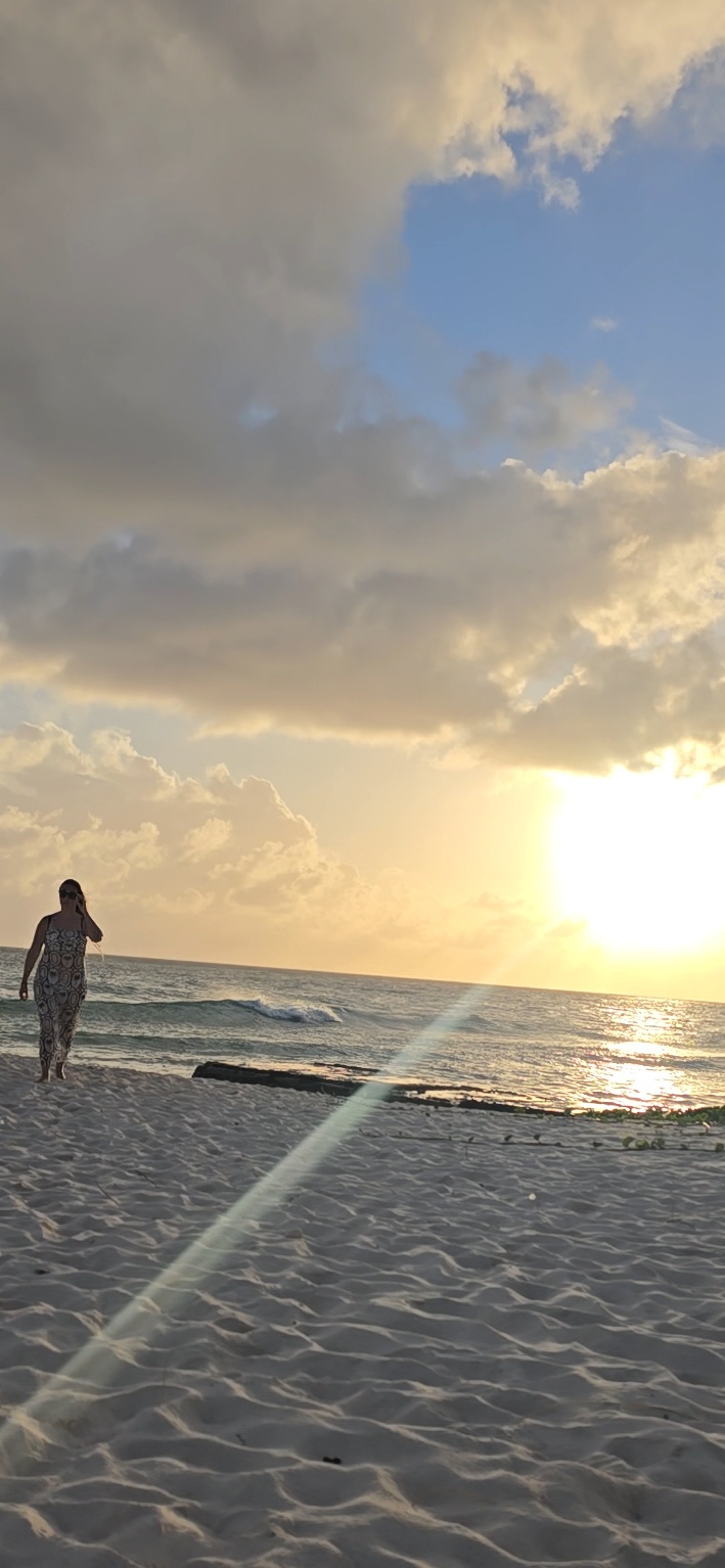 Nikki Bodie walking along a white-sand Barbados beach toward the water as the sun sets through the clouds.