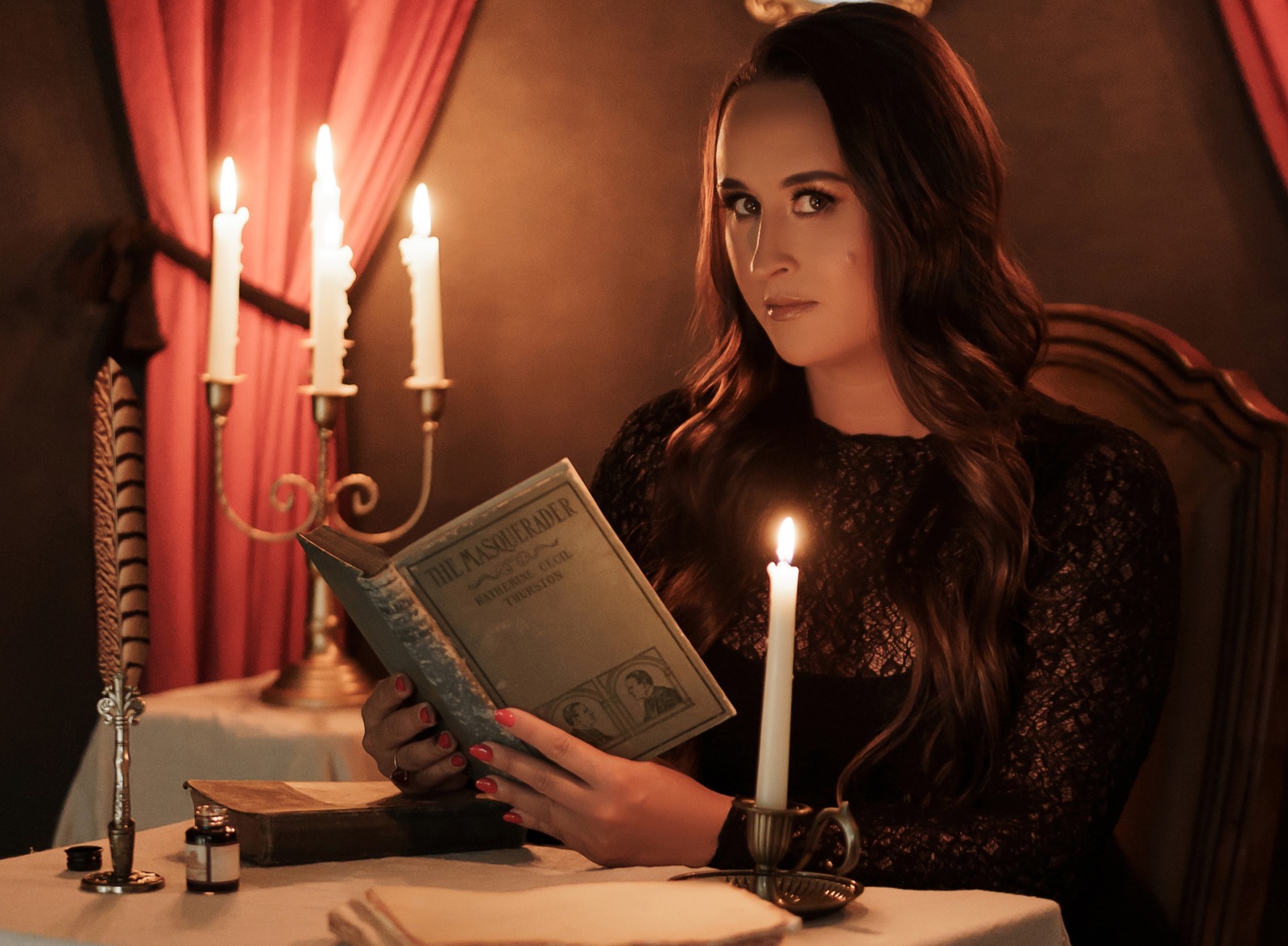 Nikki Bodie reading an antique copy of The Masquerader by candlelight at a draped table.