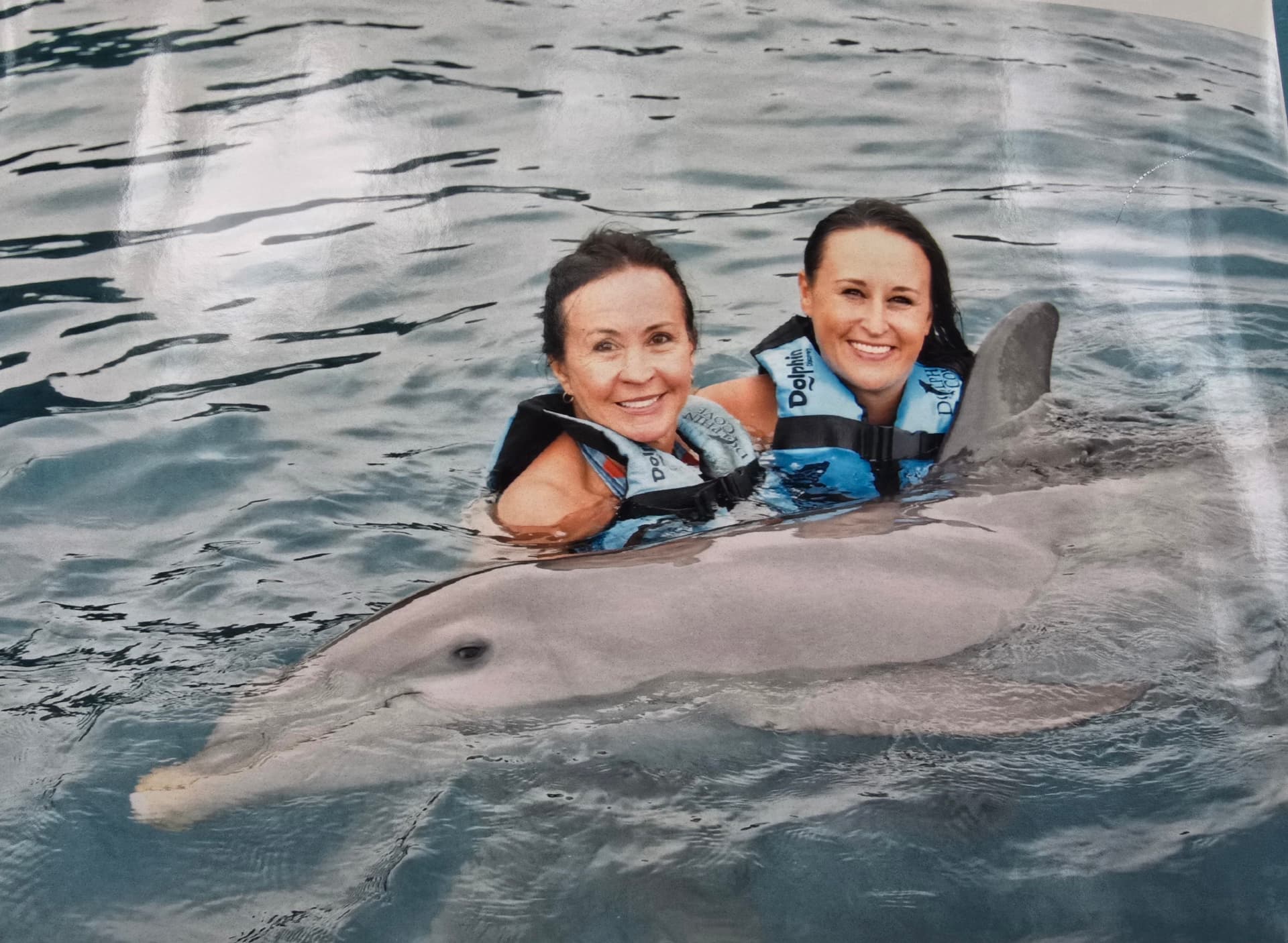 Nikki and Patti in life vests in the water, smiling beside a dolphin.