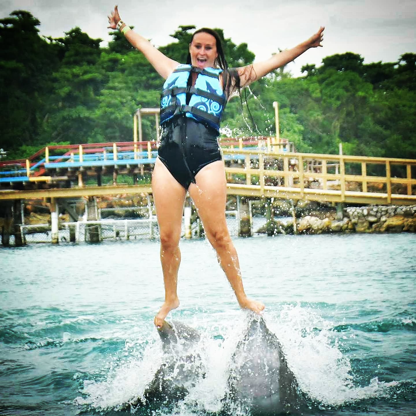 Nikki standing on the noses of two dolphins, arms up, mouth open in delight.