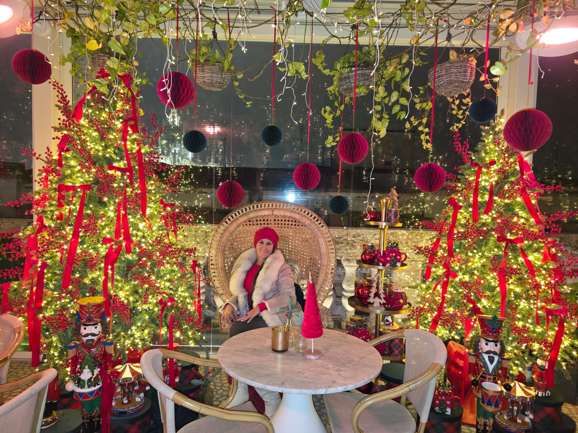 Nikki Bodie in a red hat and fur coat seated in a peacock chair between two lit Christmas trees hung with red ribbons.