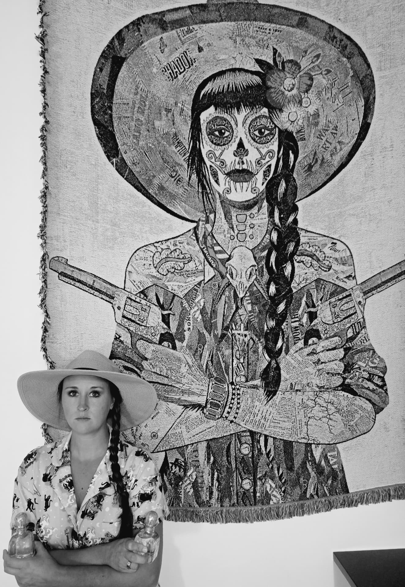 A black-and-white photo of Nikki in a wide hat and braid, arms crossed, beneath a tapestry of a woman who looks just like her.