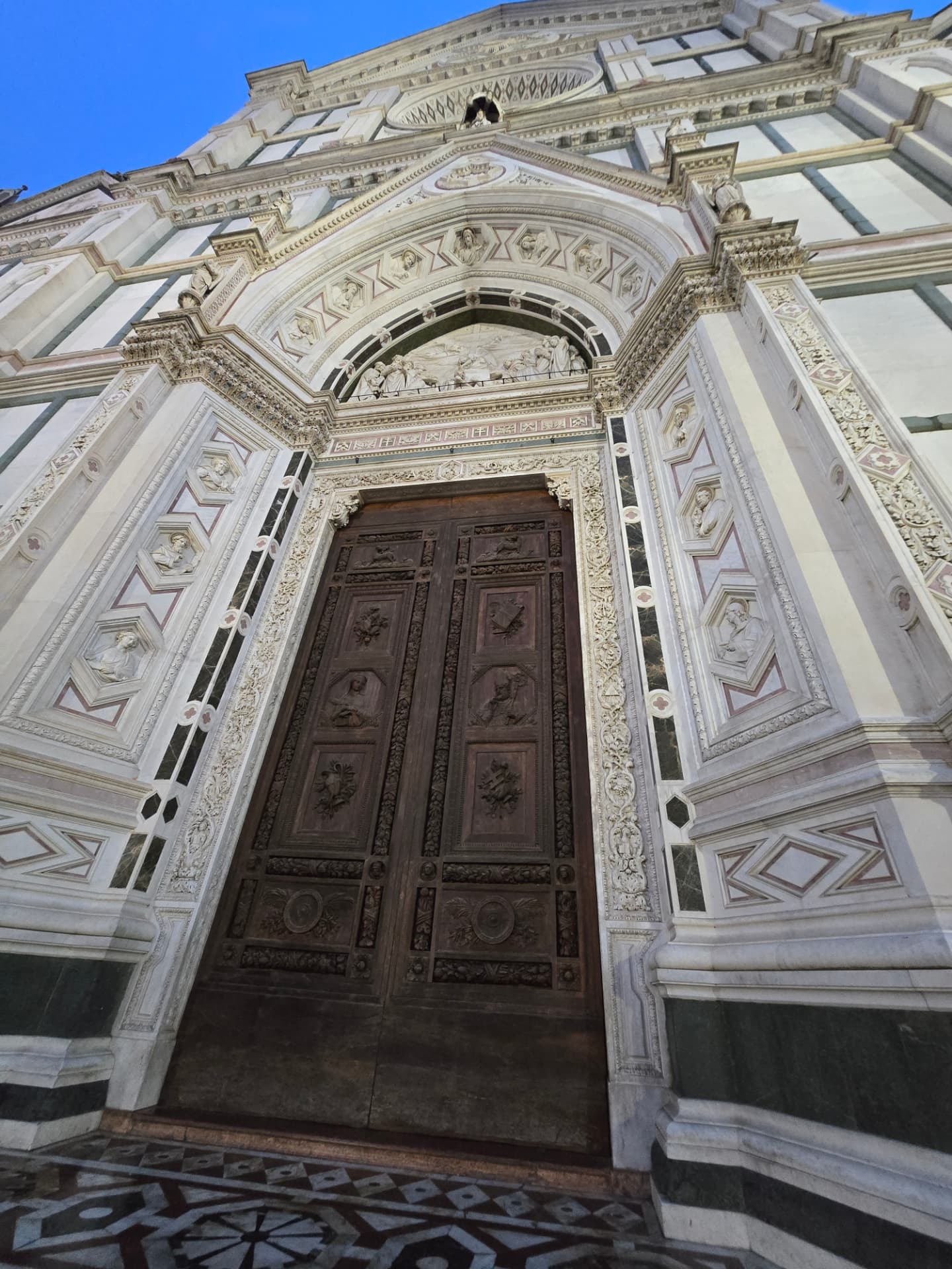 Looking up at a carved wooden door set into an ornate marble church façade in Florence.