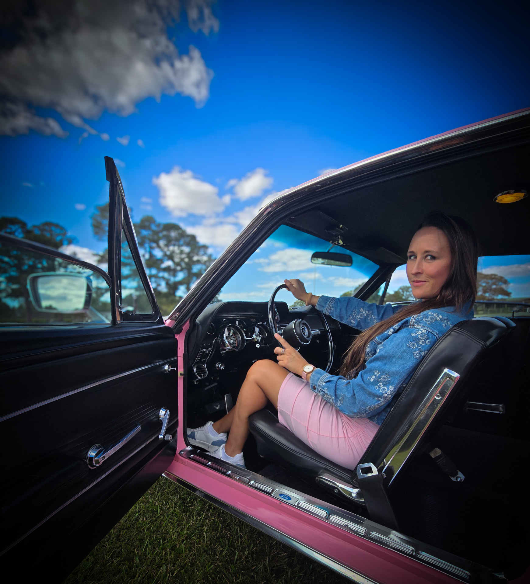 Nikki Bodie in a denim jacket sitting behind the wheel of a pink vintage Mustang with the door open.