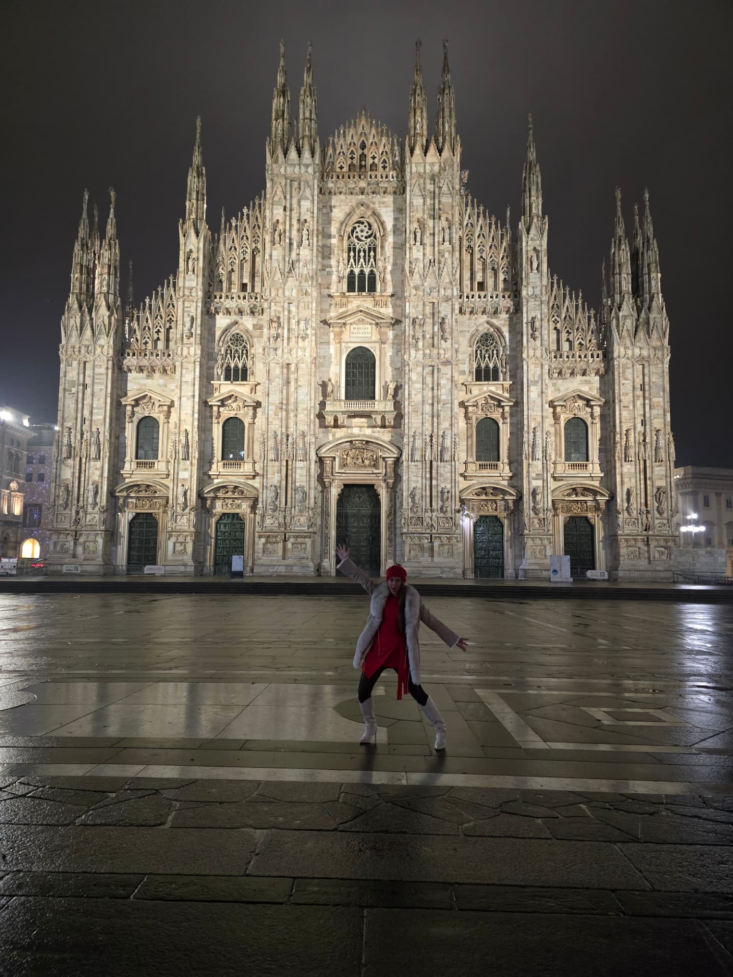 Nikki Bodie in a red coat and white boots, alone on the wet piazza before the illuminated Duomo di Milano at night.