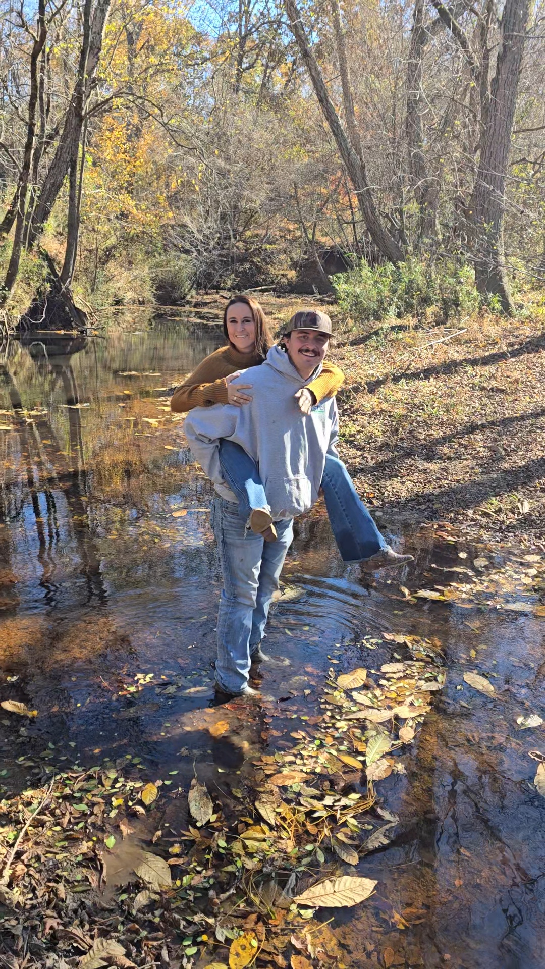 Nikki Bodie's brother carrying her piggyback through the shallow creek, both smiling, fall woods behind them.
