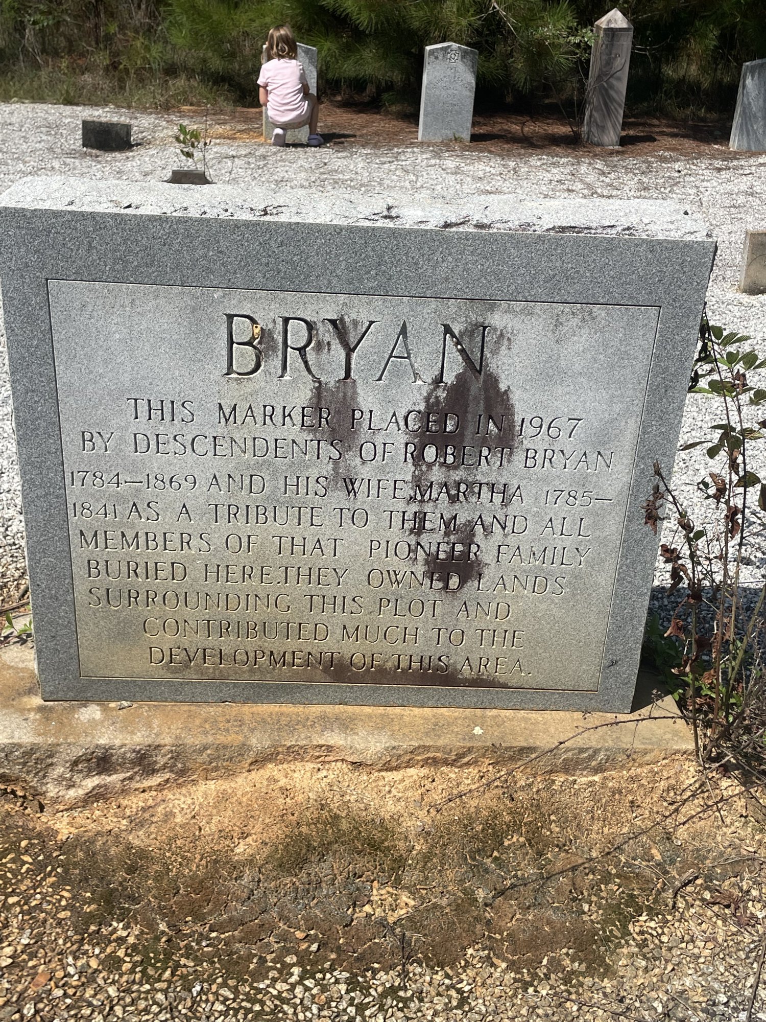 A granite marker reading BRYAN, placed in 1967 by descendants of Robert Bryan, 1784-1869, and his wife Martha, 1785-1841, as a tribute to the pioneer family buried there; older headstones and a small child stand in the gravel beyond it.