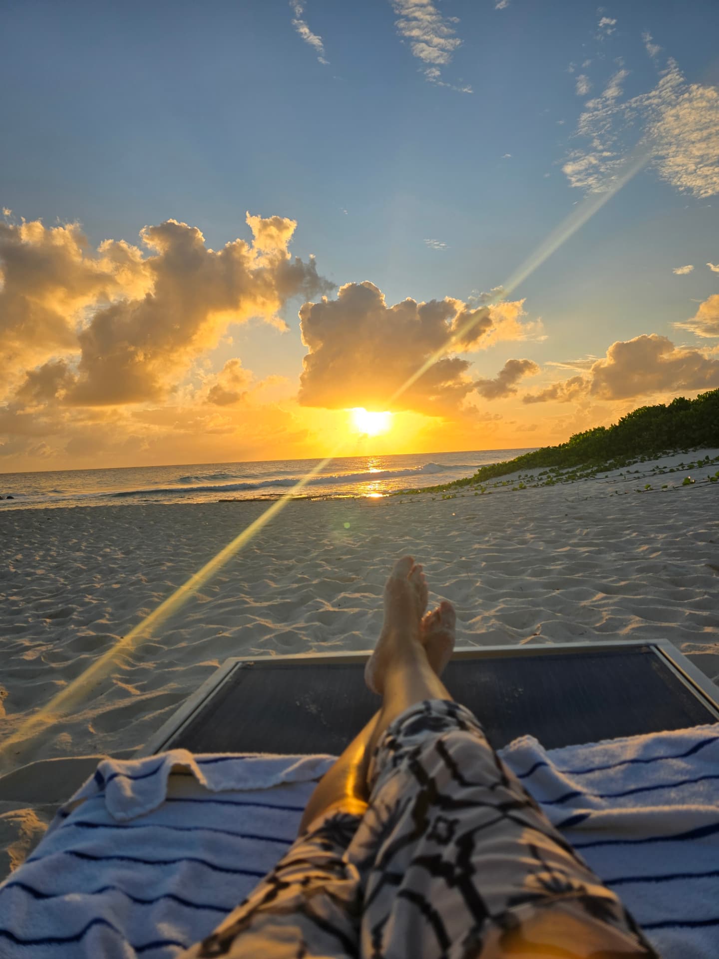 Bare crossed feet on a beach lounger facing the sun setting over the ocean in Barbados.