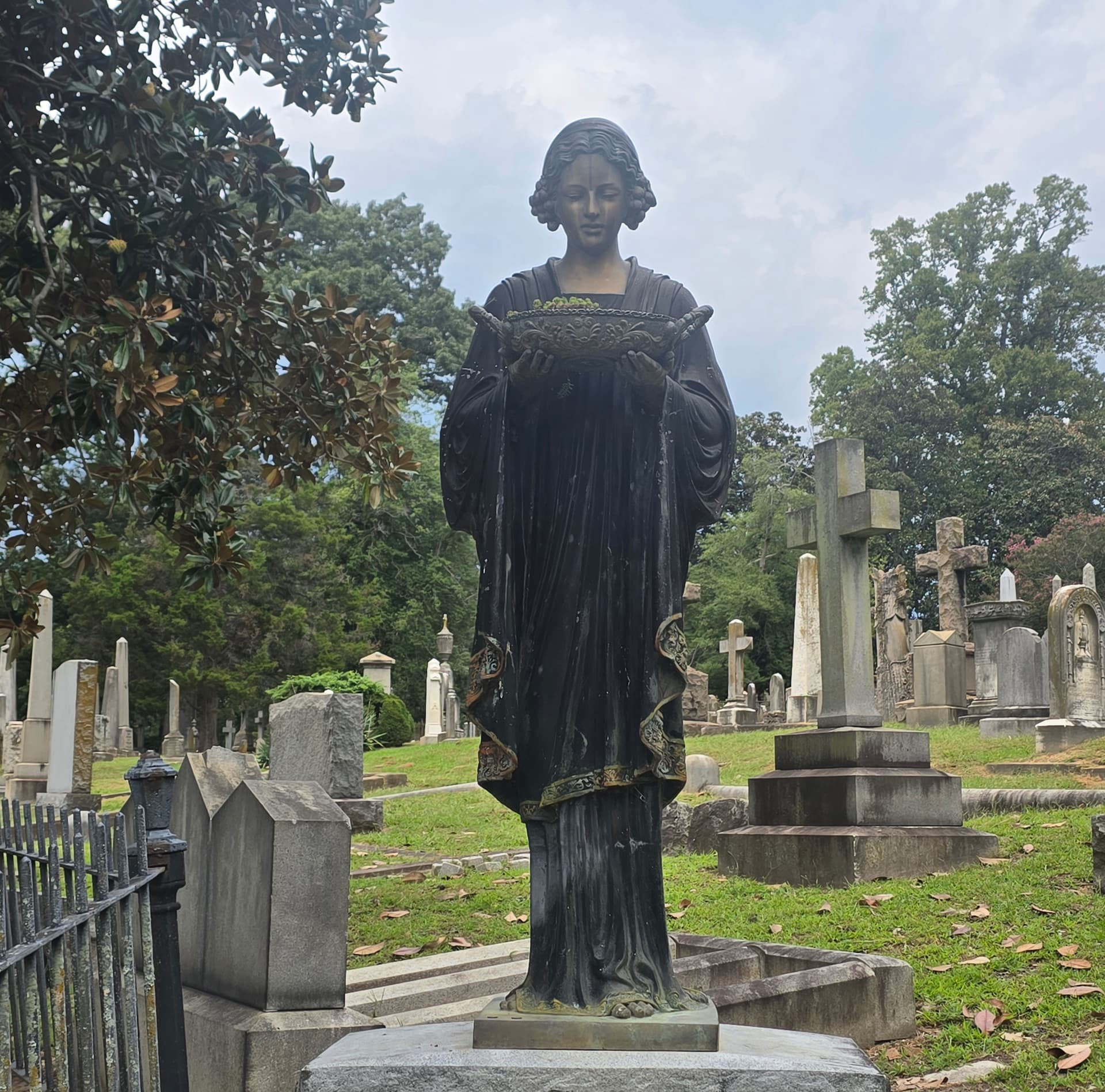 A dark bronze statue of a robed woman holding a shallow ornamented bowl, standing among crosses and headstones in Hollywood Cemetery.