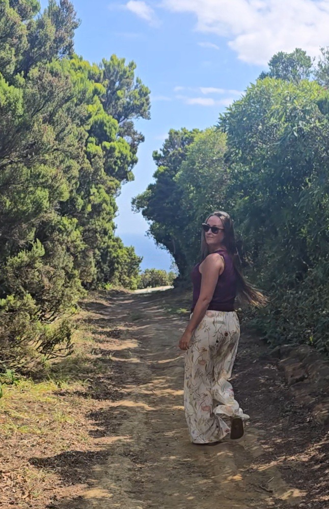 Nikki Bodie looking back over her shoulder on a sunlit dirt trail lined with green trees, the sea beyond.