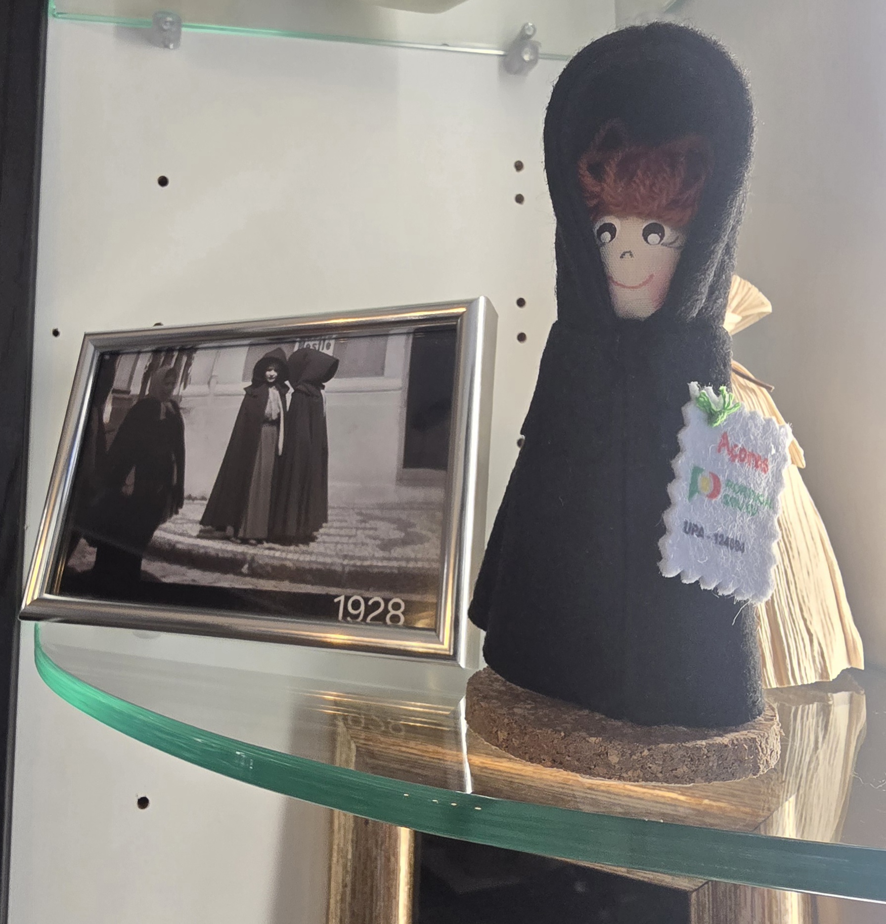 A Mulher de Capote doll in a black hooded cloak beside a framed 1928 photograph of cloaked Azorean women.