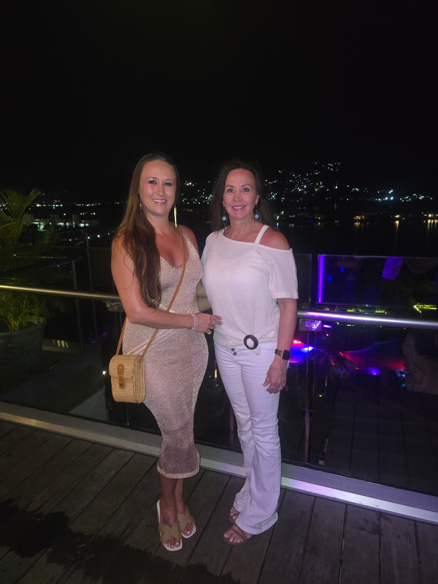Nikki and Patti dressed up at night on a rooftop terrace, the lights of the Jamaican coast behind them.