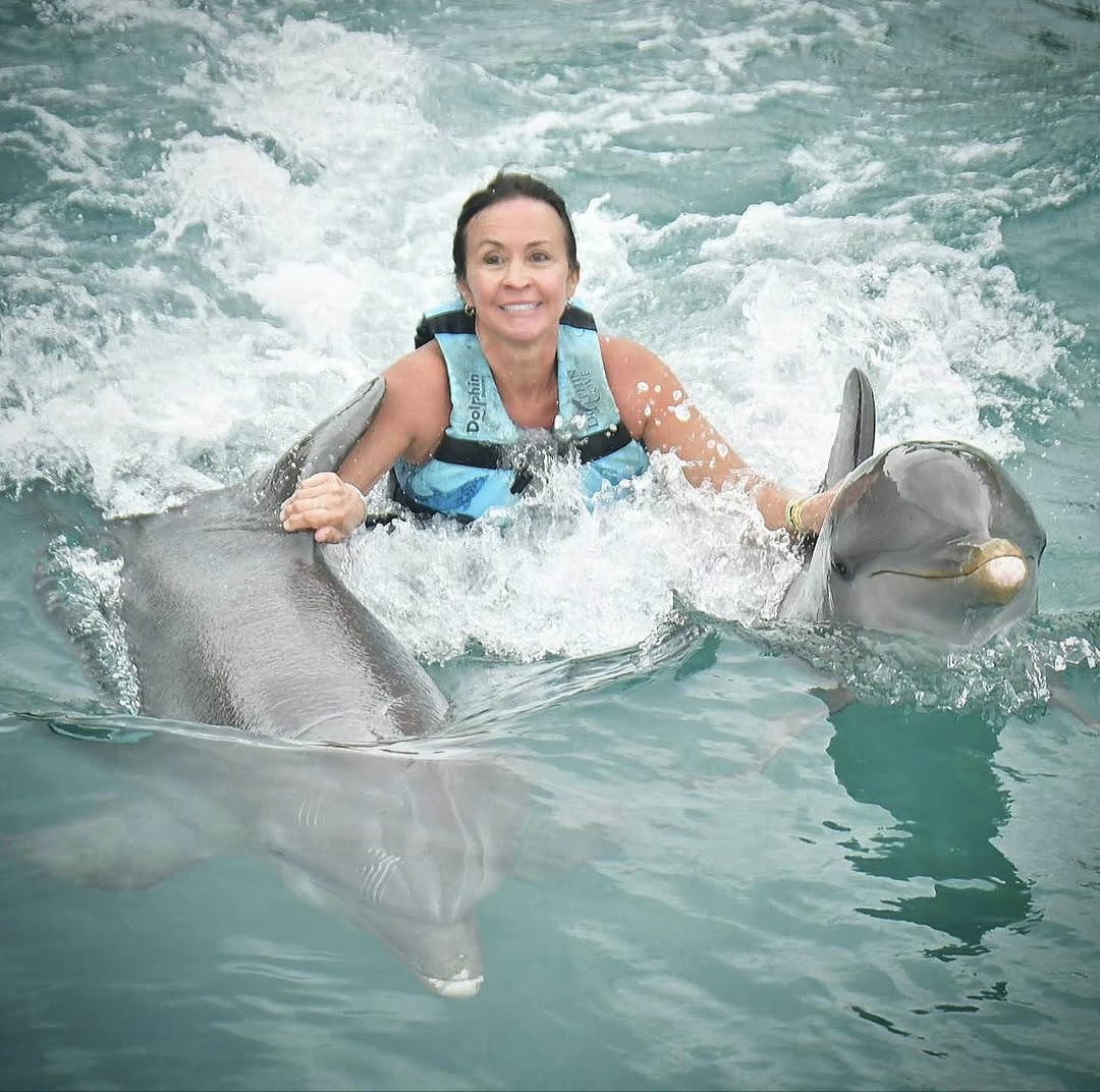 Patti in a life vest pulled through the water between two dolphins, holding their fins and smiling.