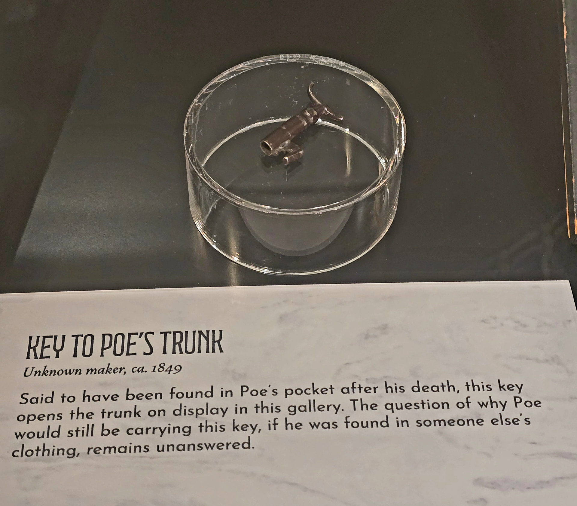 A small iron key in a glass dish in a museum case, above a label reading "Key to Poe's Trunk, unknown maker, ca. 1849."