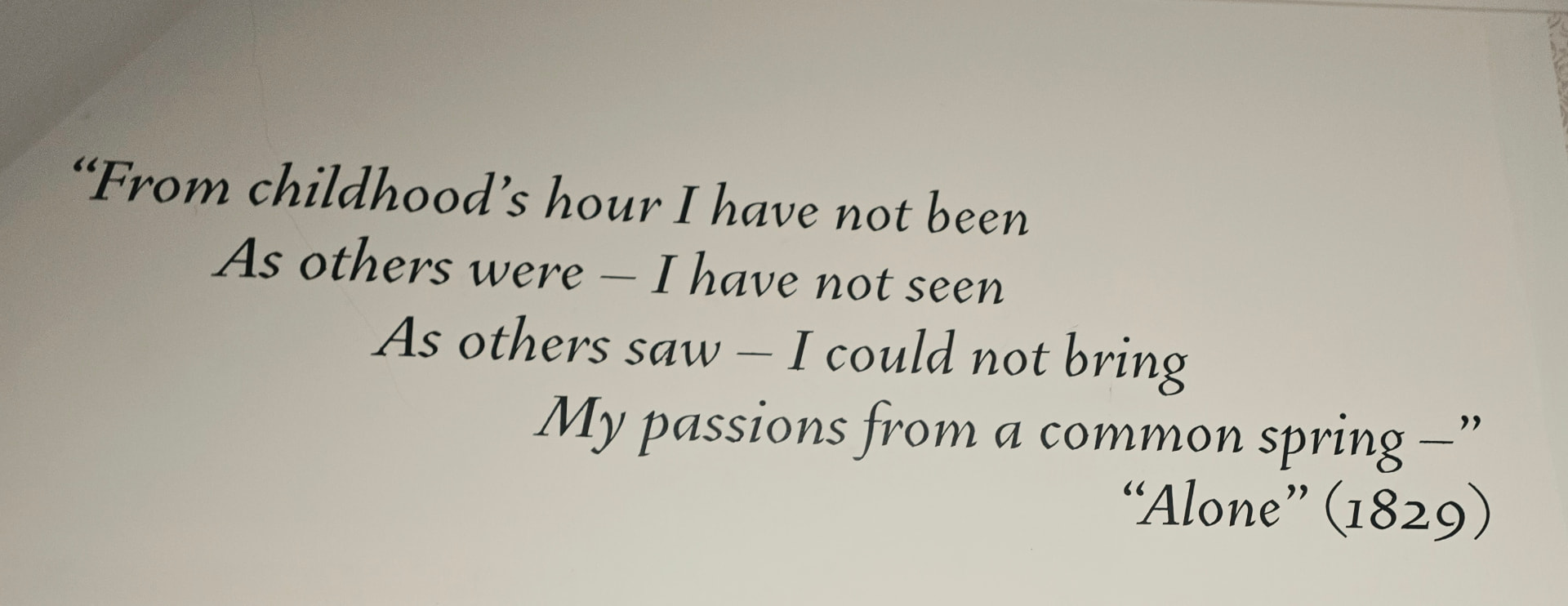 A museum wall printed in italic type with the opening lines of Poe's poem "Alone" (1829): "From childhood's hour I have not been as others were."