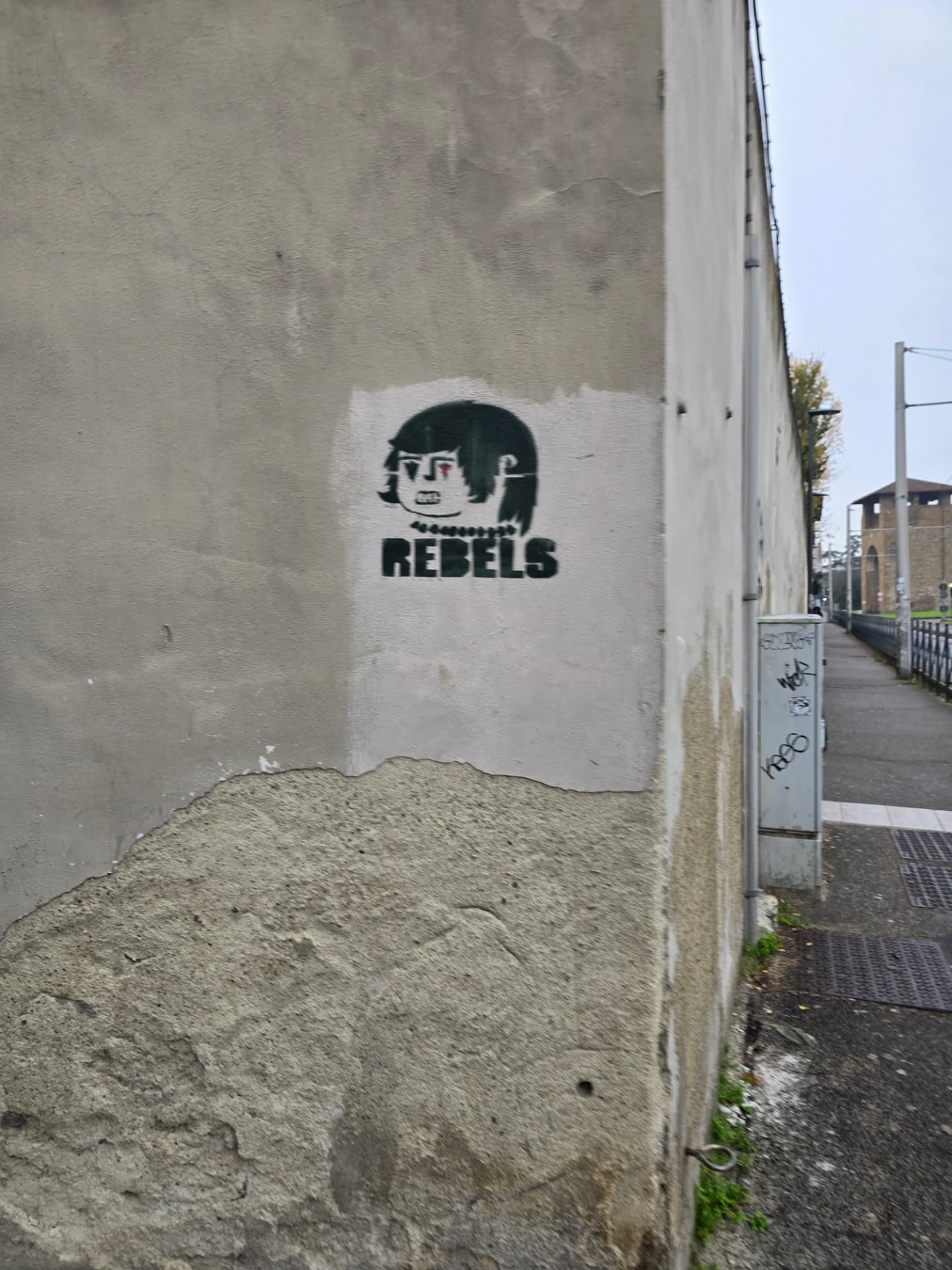 A black stencil of a dark-haired face above the word REBELS on a weathered wall beside a long street.