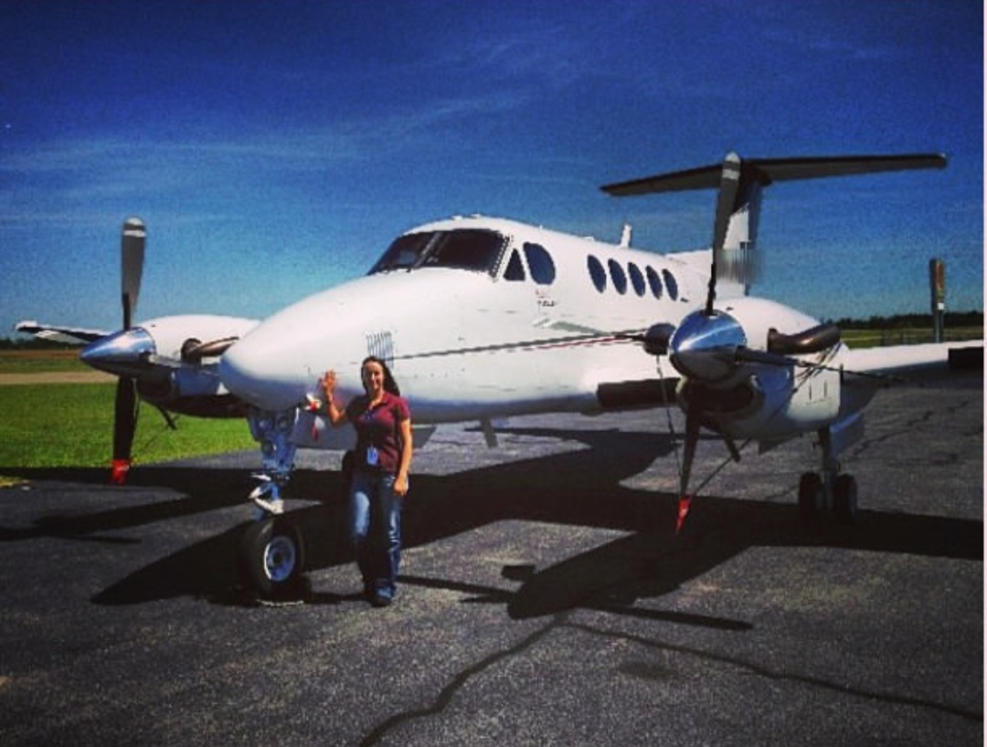 Nikki waving beside a King Air on a sunny ramp.