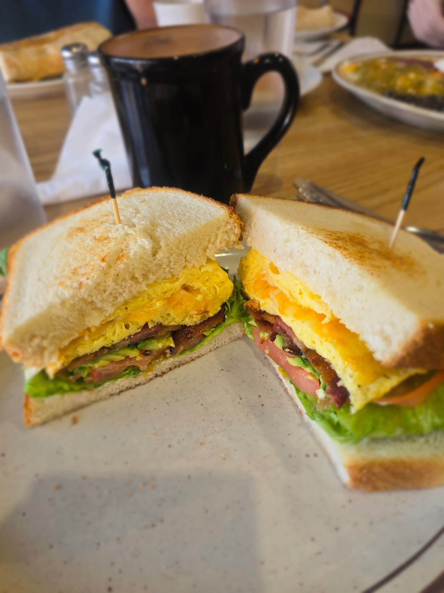 A breakfast sandwich with egg, bacon and avocado beside a black mug of coffee.