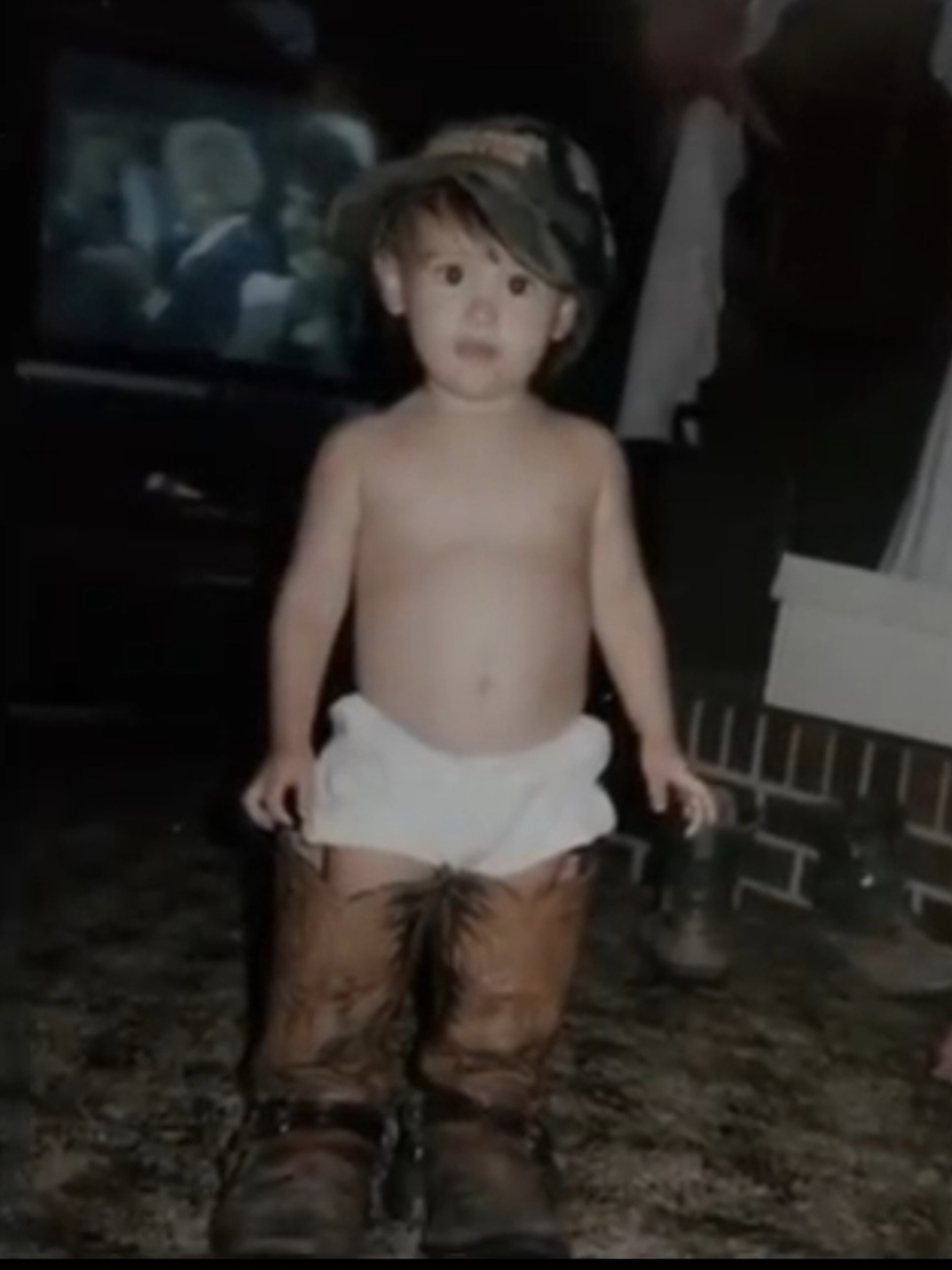 Nikki Bodie as a toddler in South Carolina, standing in a pair of grown-up cowboy boots and a cap.
