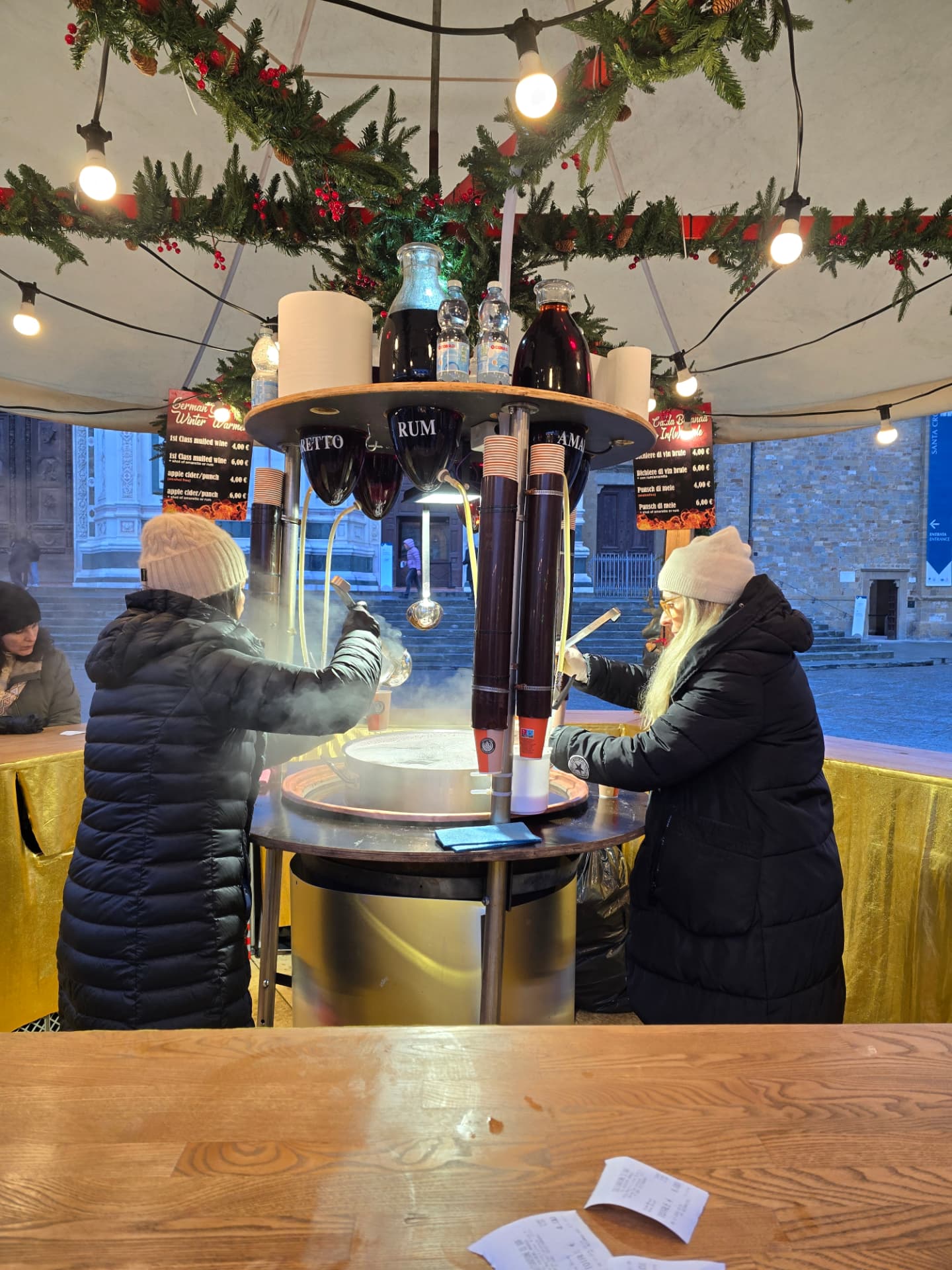 A round wooden Christmas market bar hung with garland, people ordering drinks.