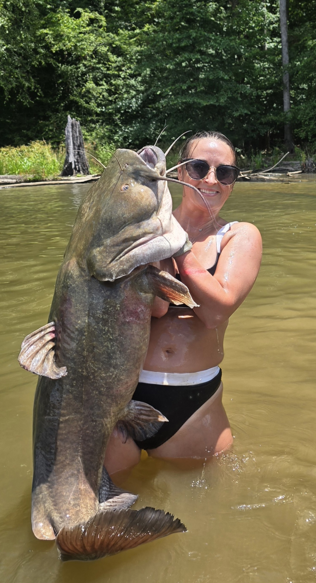 Nikki standing in the river holding a catfish nearly as tall as she is.