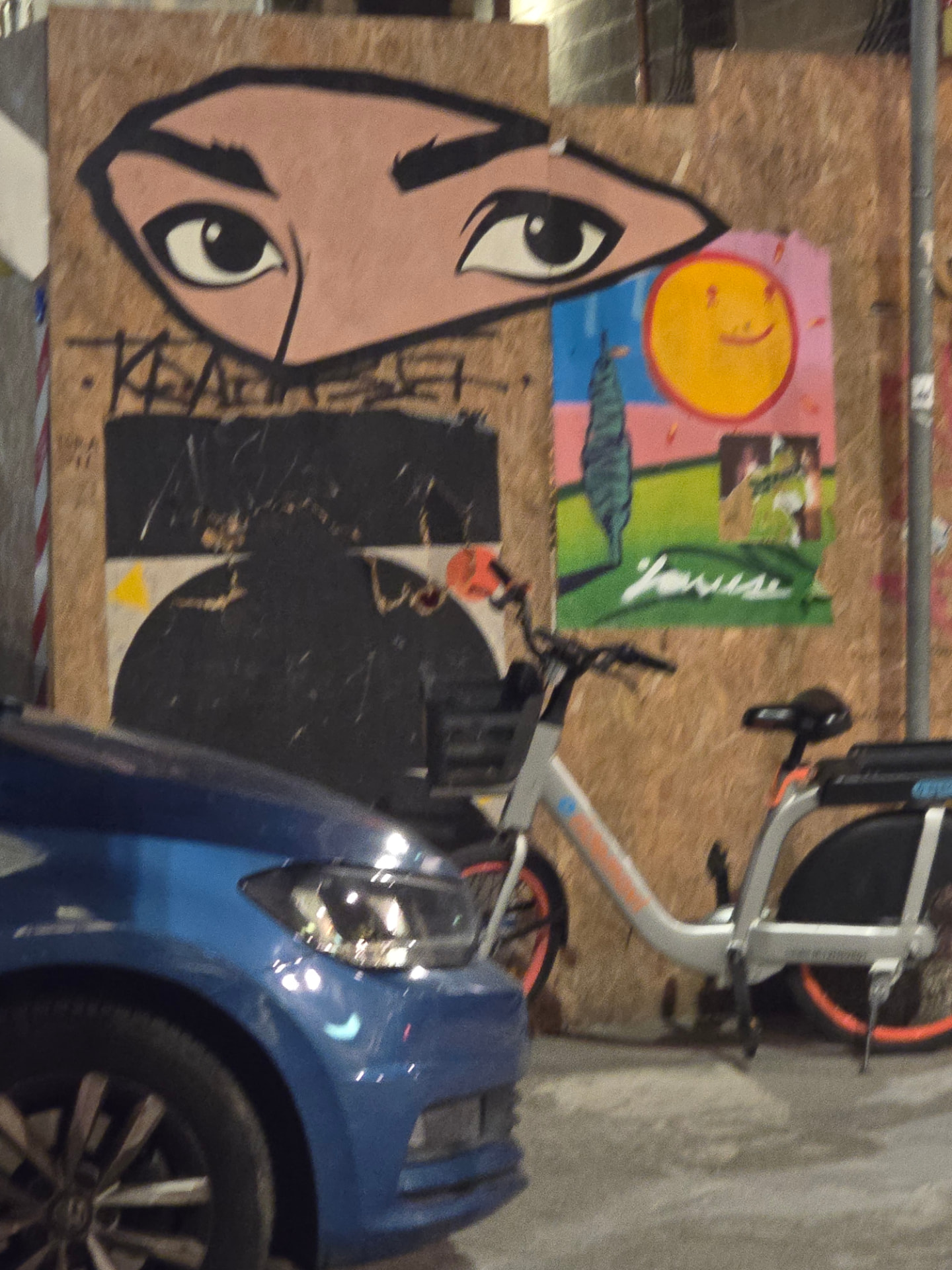 A mural of large watchful eyes and a smiling sun on plywood hoarding, a bike and blue car in front.
