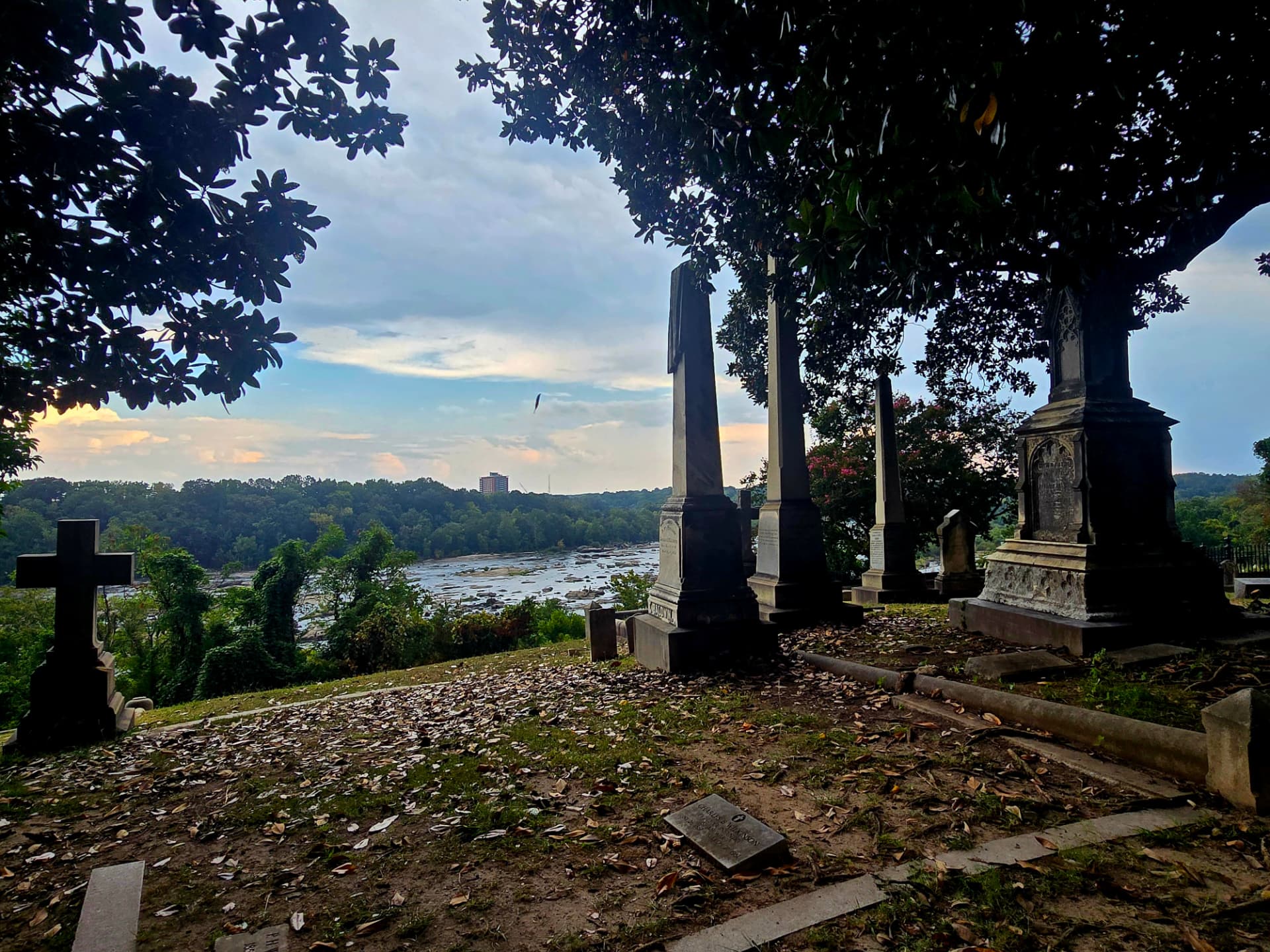 Dusk over the James River seen from the hillside of Hollywood Cemetery, obelisks and a stone cross silhouetted beneath magnolia branches as clouds gather.