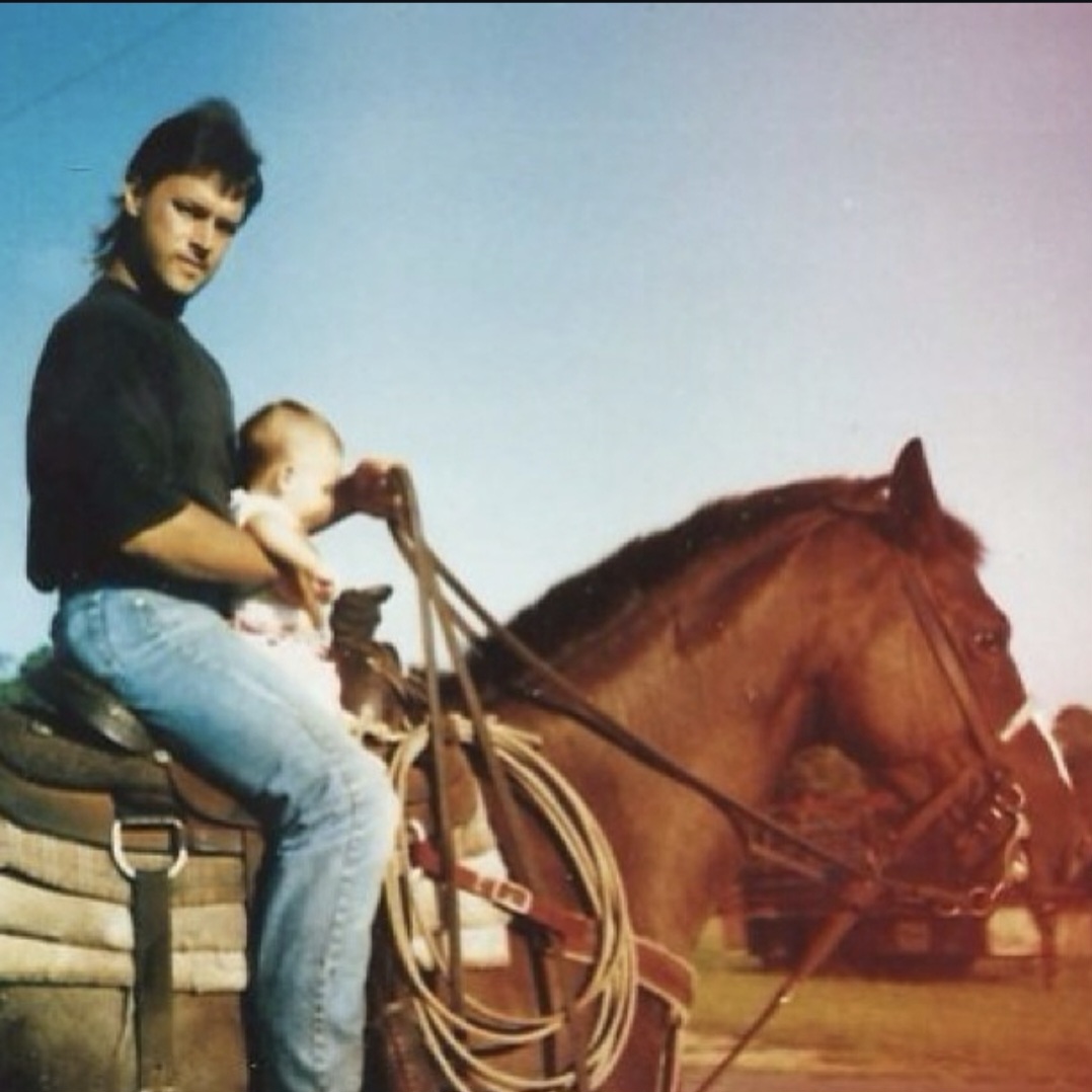 Nikki Bodie as a baby held on horseback by her father, a rope coiled on the saddle.