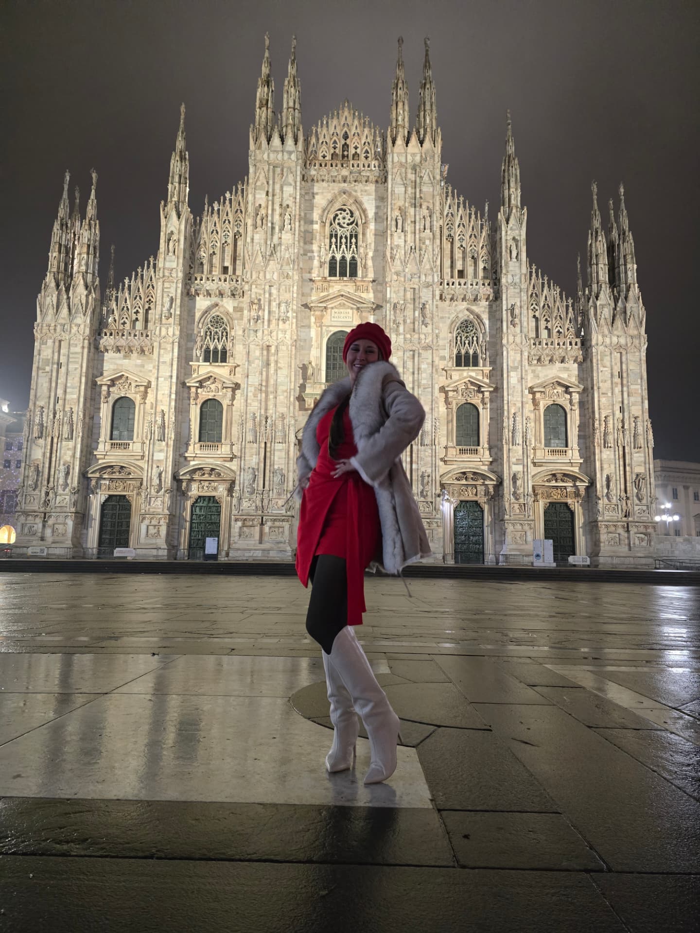 Nikki Bodie in a red coat and fur stole standing before the Duomo di Milano at night on wet stone.