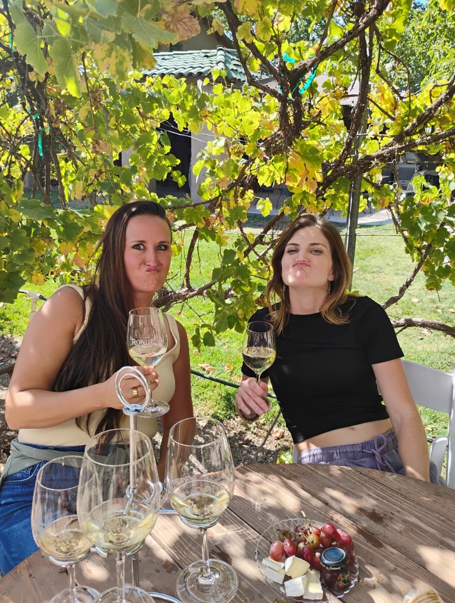 Nikki and Ali at a winery table under the vines with glasses of white wine.