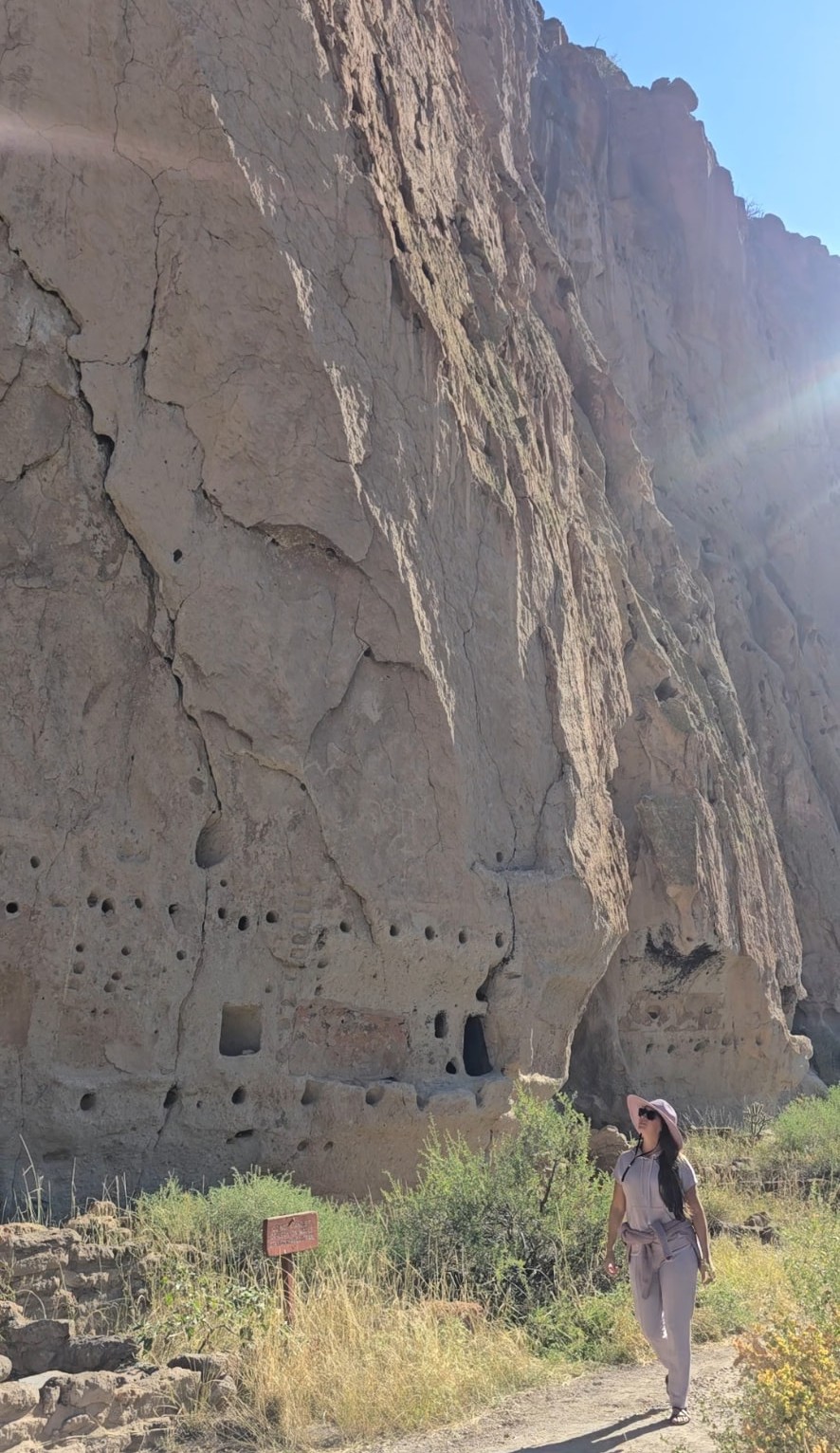 A wide view of the pocked cliff wall in Frijoles Canyon, Nikki small on the trail below.