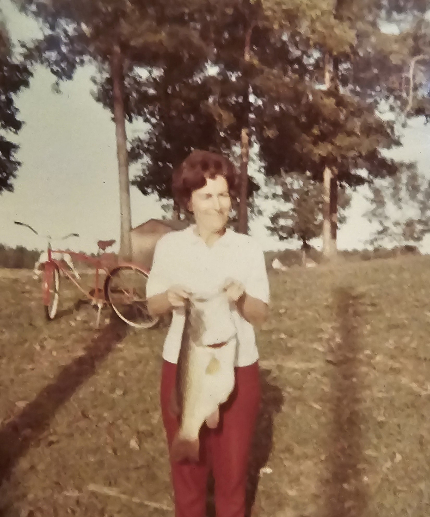 A faded color photograph of Grandma Reba holding a large bass, a red bicycle behind her.
