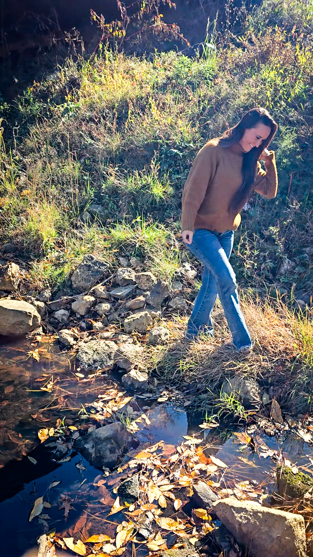 Nikki Bodie stepping along the rocky bank of a South Carolina creek in fall sunlight, leaves floating on the dark water.