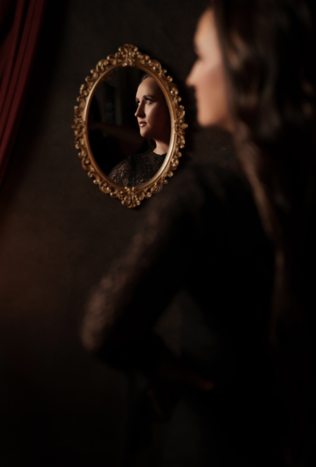 Nikki Bodie's profile reflected in an ornate gold oval mirror on a dark wall, her blurred figure in the foreground.
