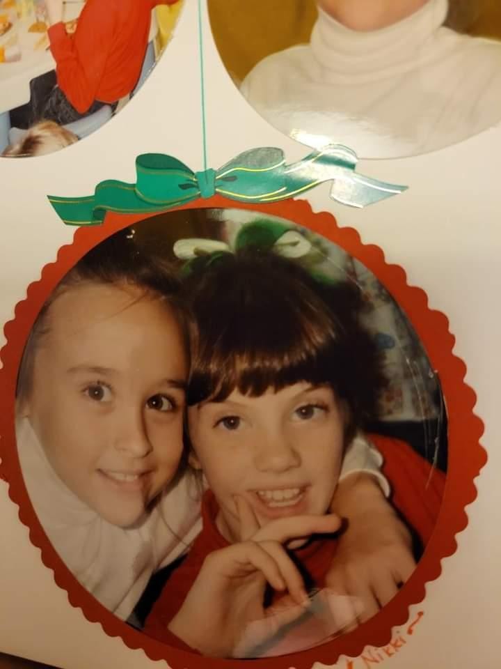 A childhood photo of Nikki and Ali, cheek to cheek, set in a round red ornament frame.