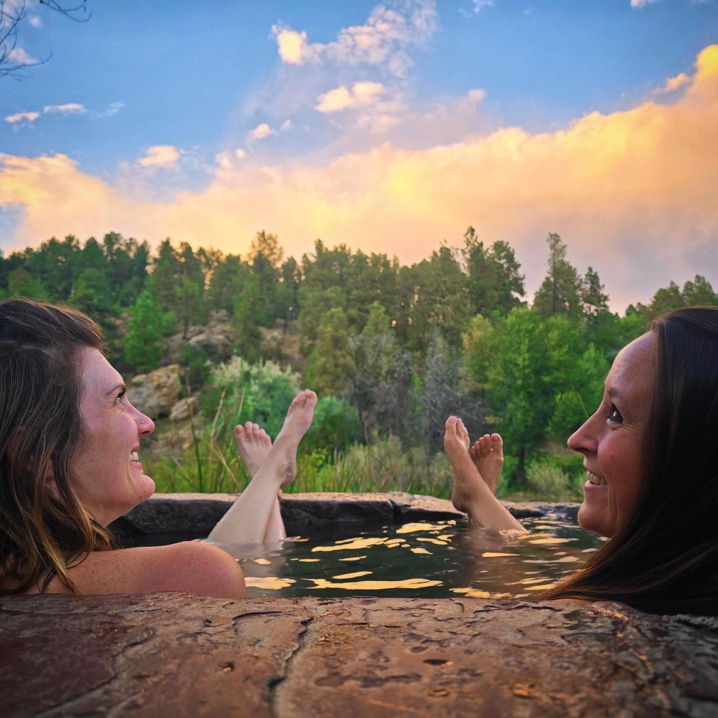 Nikki and Ali in an outdoor hot spring at sunset, smiling at each other with their feet up.