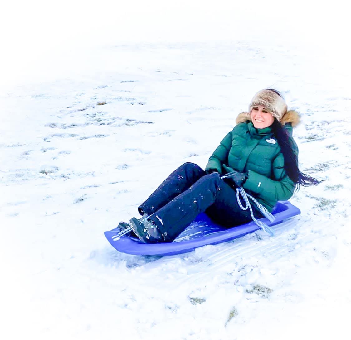 Nikki Bodie in a green coat and fur hat smiling on a blue sled in the snow.