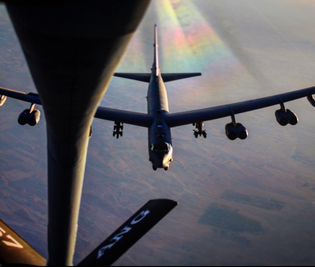 A B-52 behind the refueling boom, a rainbow on the ground below.