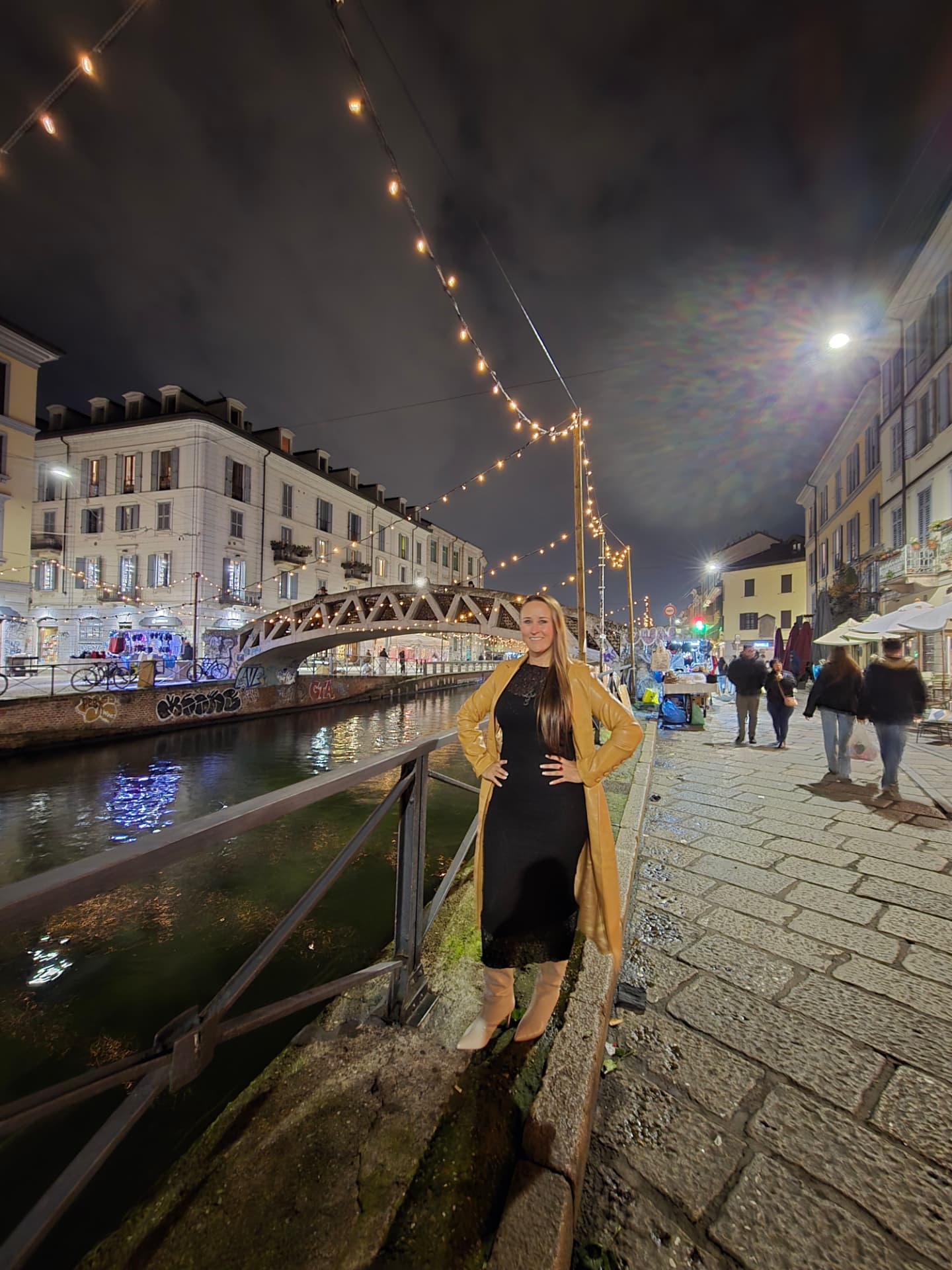 Nikki Bodie leaning on a railing beside a Milan canal at night, string lights and a footbridge behind her.