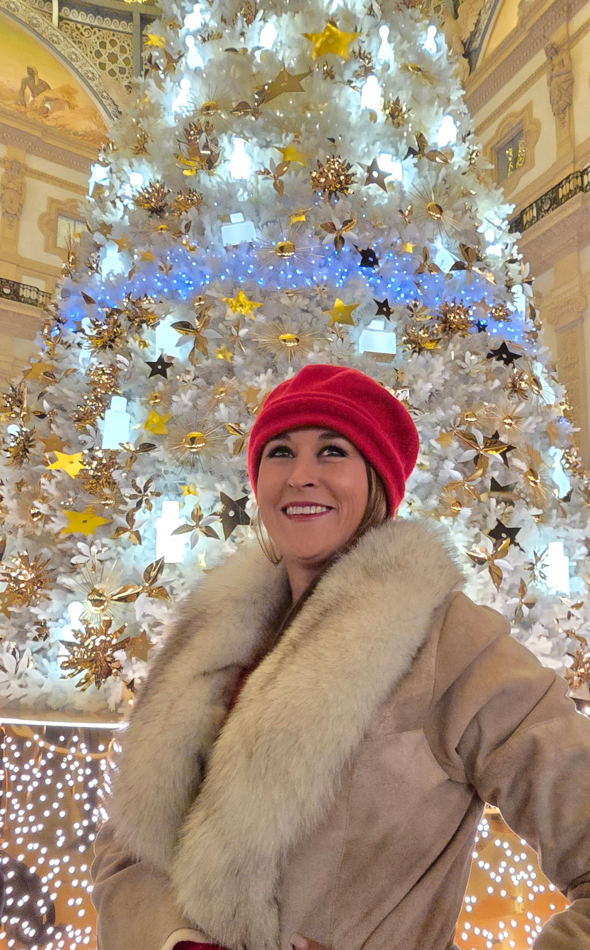 Nikki Bodie in a red beret smiling beneath an enormous white and gold Christmas tree in a domed gallery.