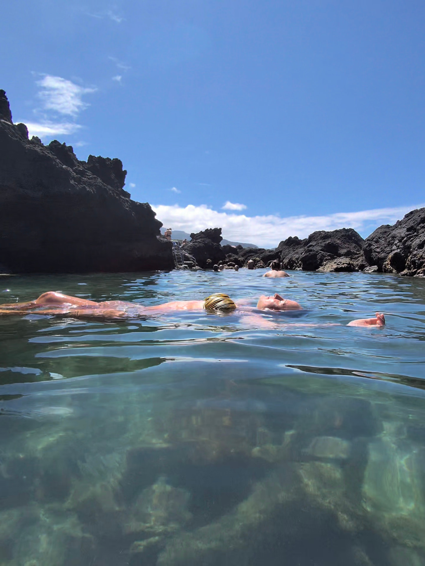 Nikki floating in a clear volcanic pool at Biscoitos, black rock all around.