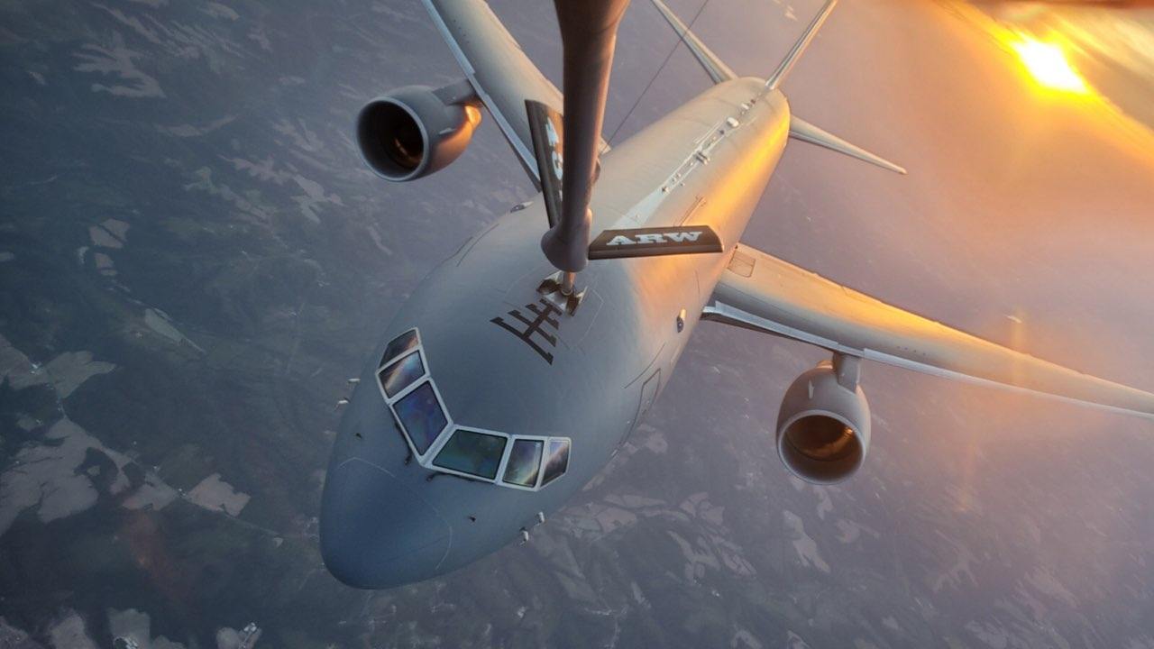 A KC-46 nosed up beneath the refueling boom at sunset, gold light across its wing.