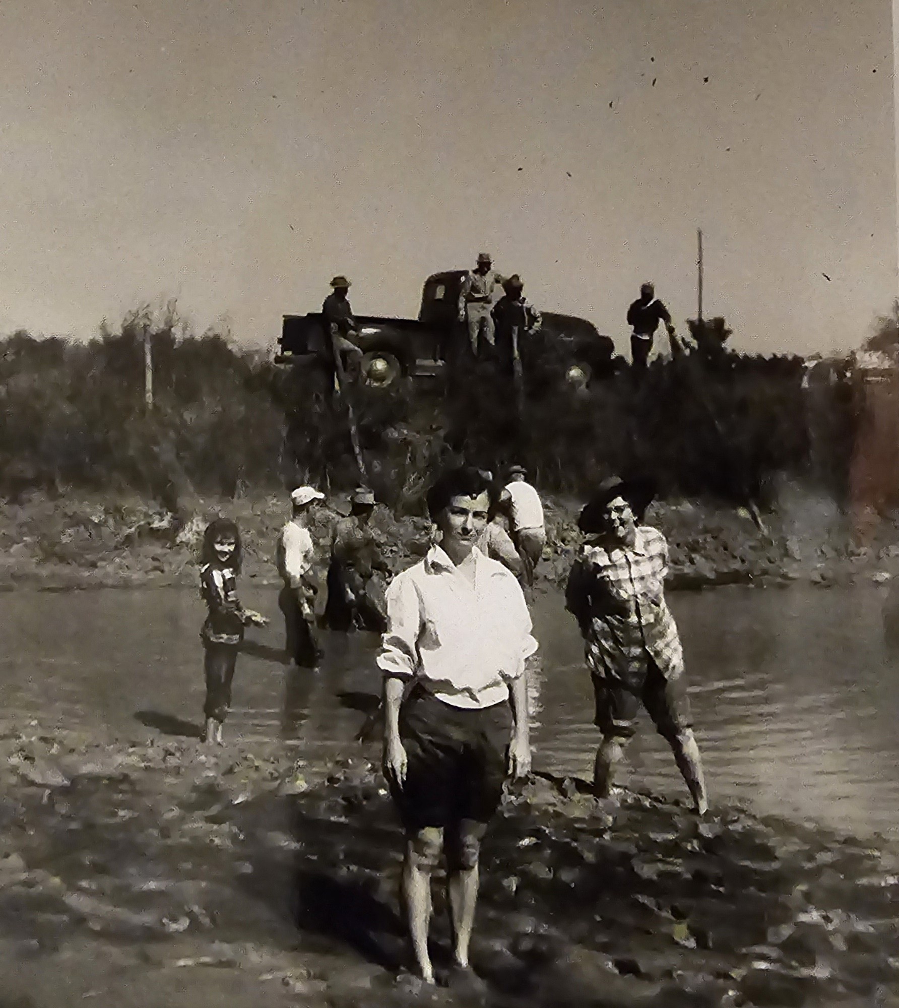 A black-and-white photograph of family wading in a shallow pond, men seated on an old truck on the bank behind them.